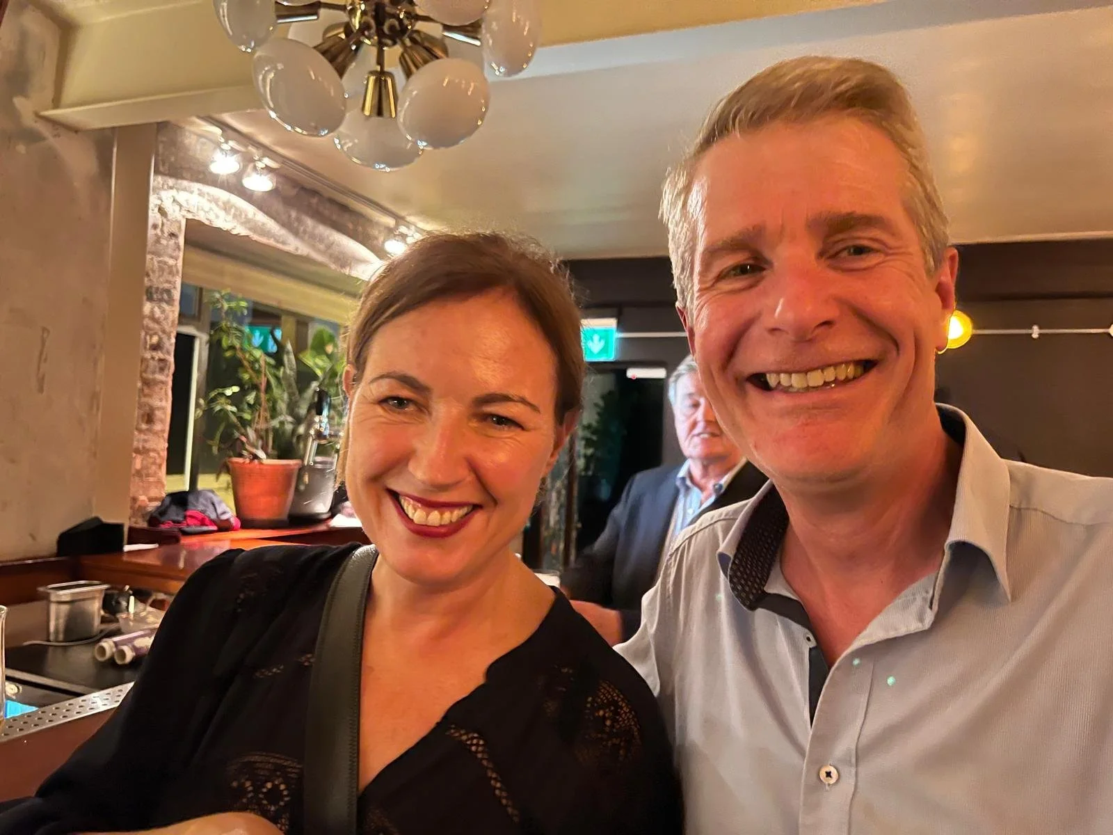 Two smiling adults, a woman with short dark hair and a man with short blonde hair, taking a selfie at a social gathering in a warmly lit indoor setting, with a third person in the background.