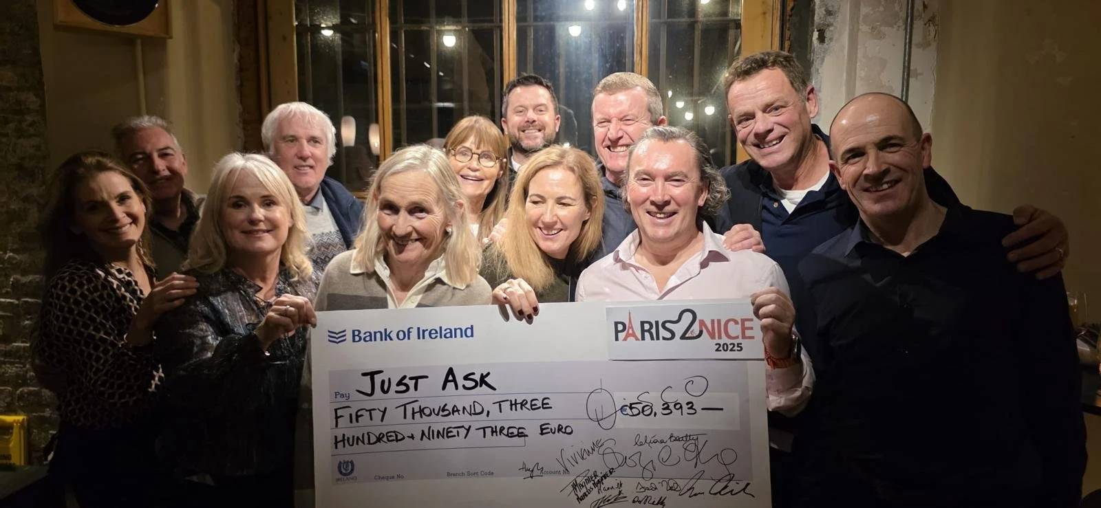 Group of people gathering around a large check with the logos of Bank of Ireland and Paris 2 Nice for 50,393 euros. They are smiling and appear to be celebrating in a warmly lit indoor setting.