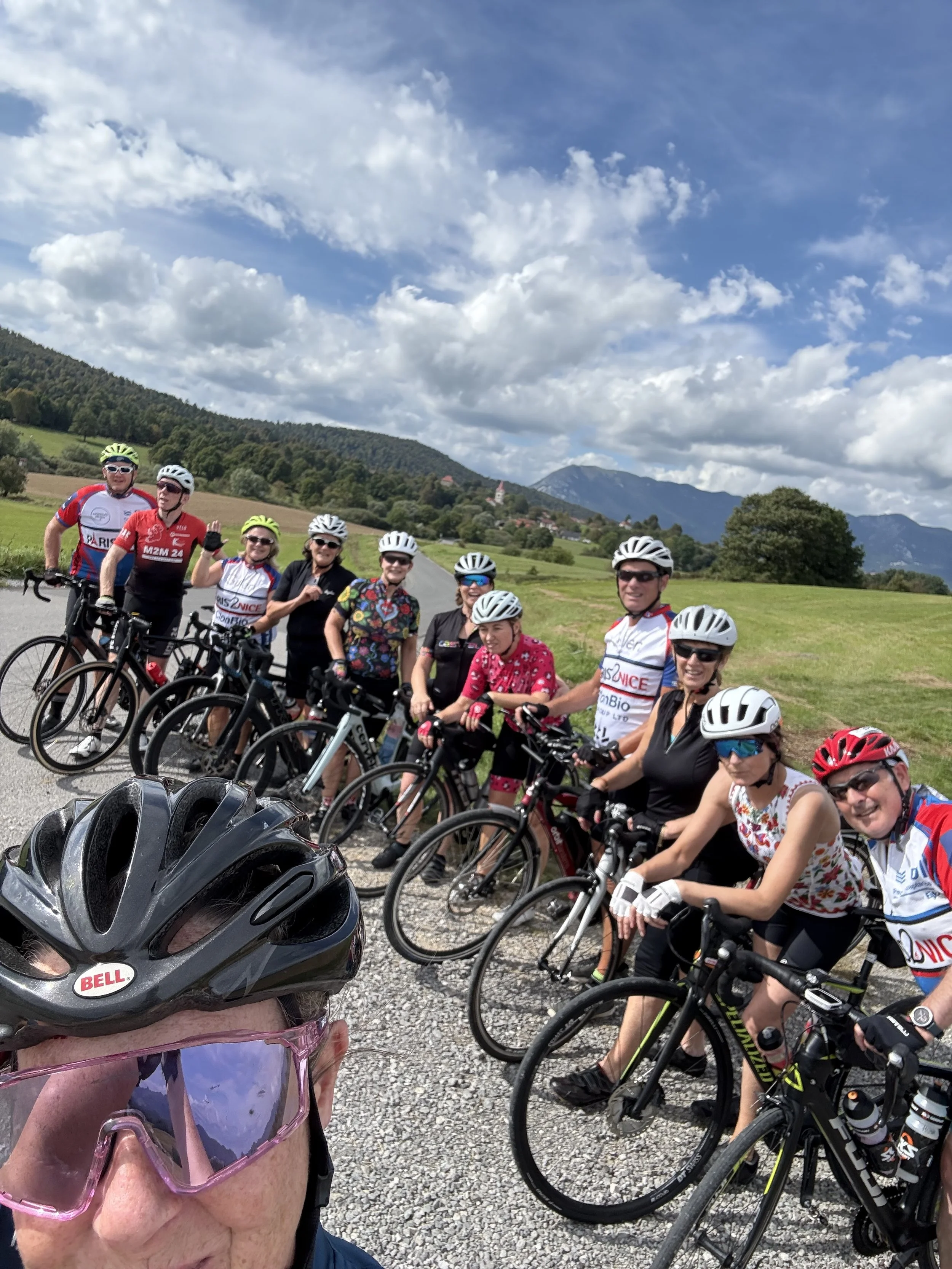 A group of people wearing cycling helmets and gear posing with their bicycles on a rural road, with green hills and mountains in the background under a partly cloudy sky.