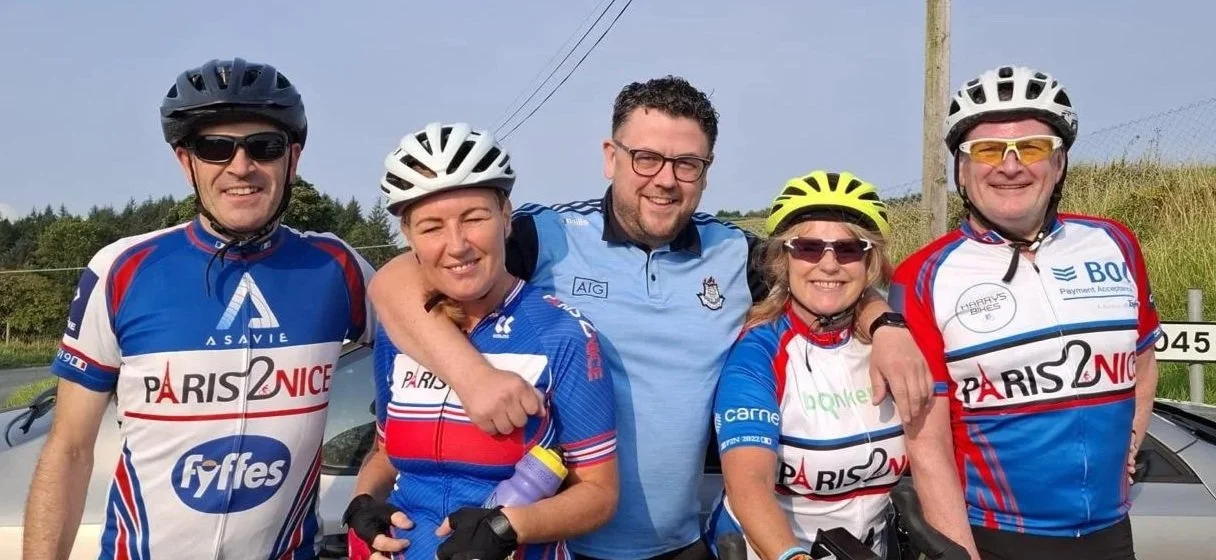 Group of five people in cycling gear, smiling and posing outdoors, wearing helmets and sunglasses.