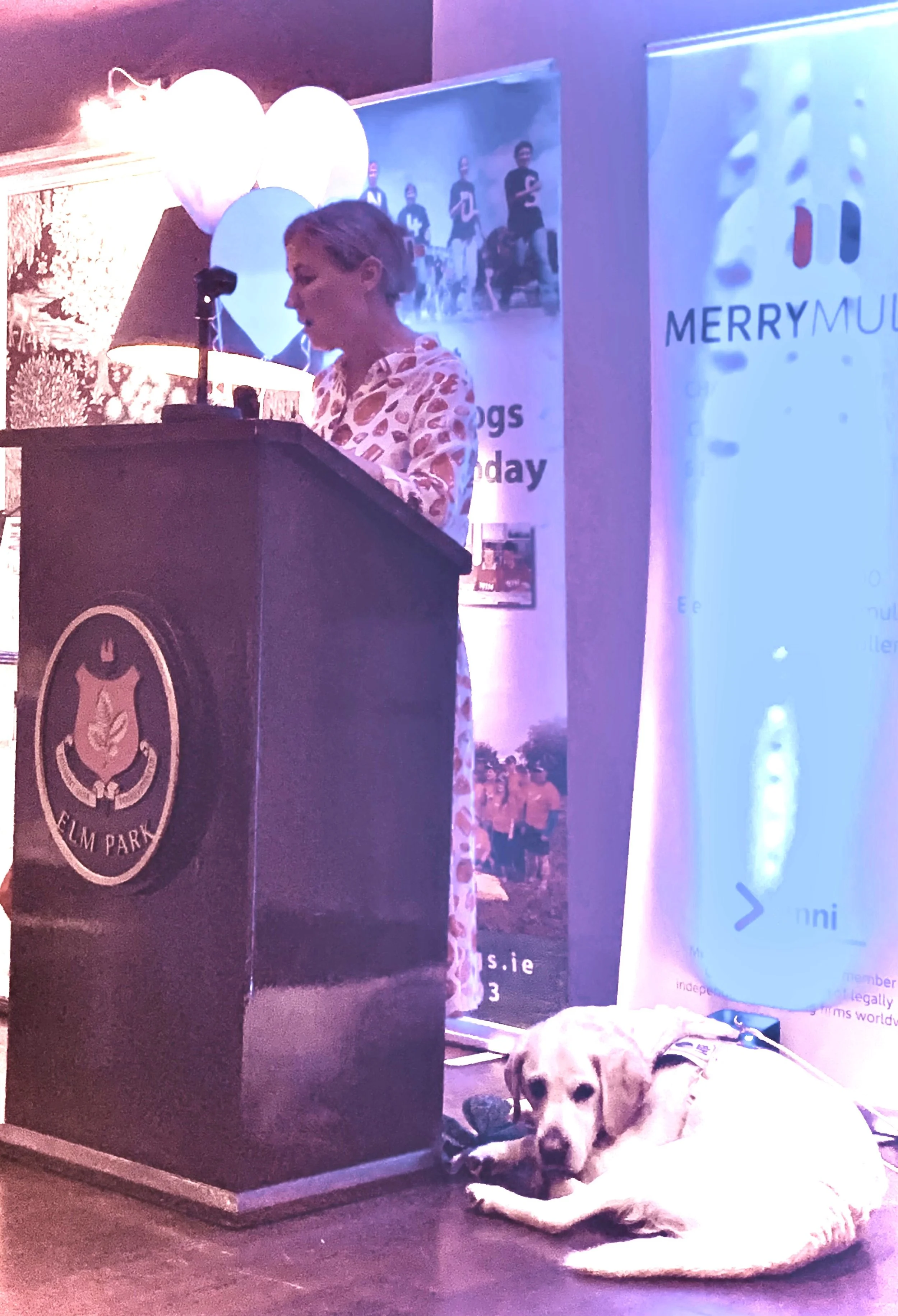 A woman in a patterned dress stands at a podium with a logo, speaking into a microphone, at a decorated event with balloons and banners, with a dog lying on the floor nearby.