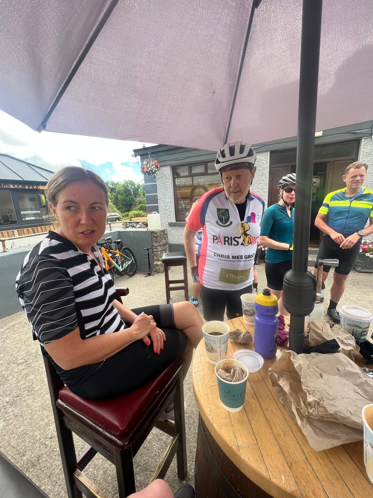 Group of people, including a woman sitting on a stool and three others standing, gather outdoors under a large umbrella, with a table holding drinks, cups, and food, after cycling.