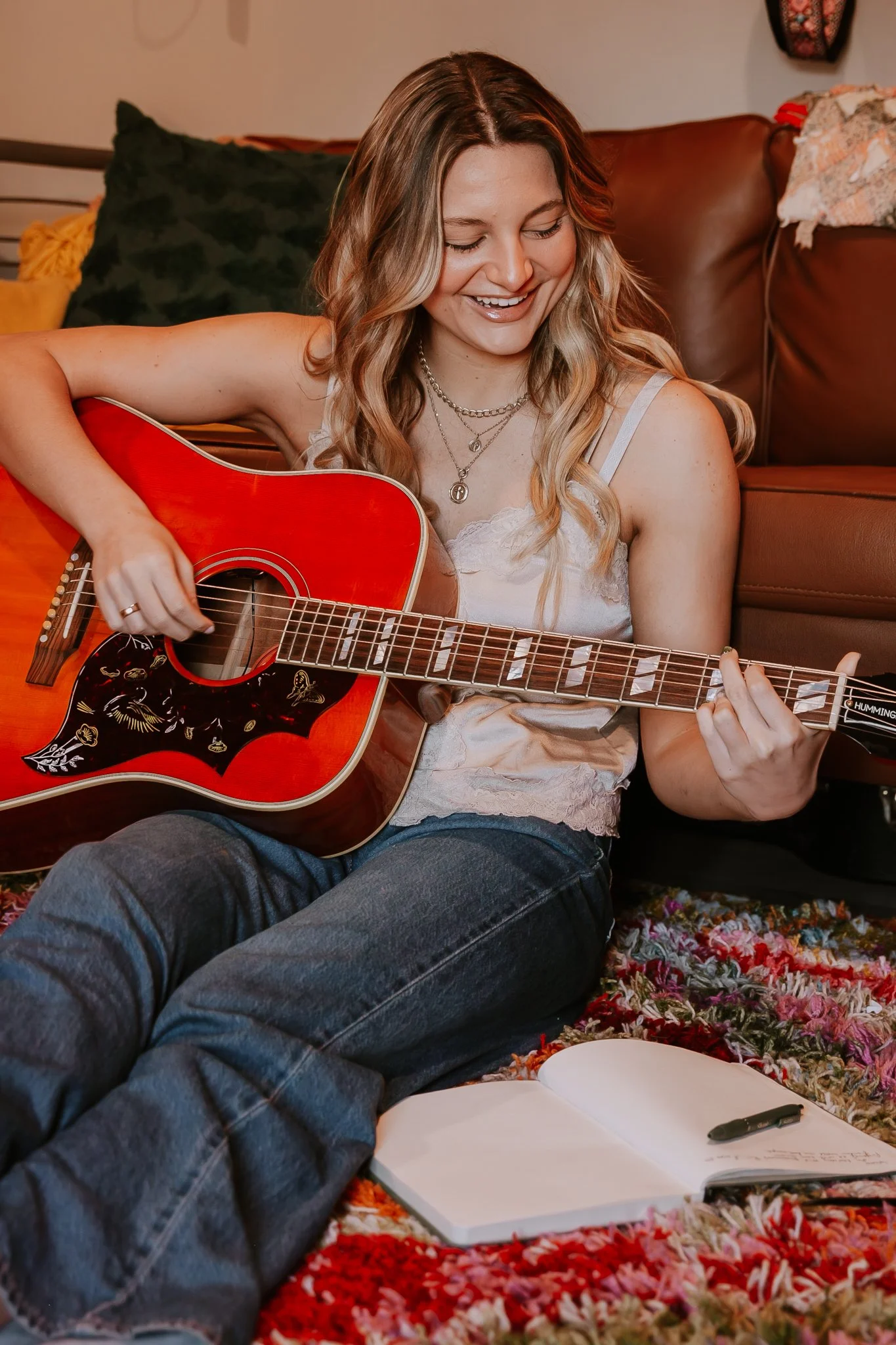 A young woman with long, wavy blonde hair sitting on a multicolored rug, playing a red acoustic guitar, smiling, with an open notebook and pen on the floor nearby, in a cozy living room.