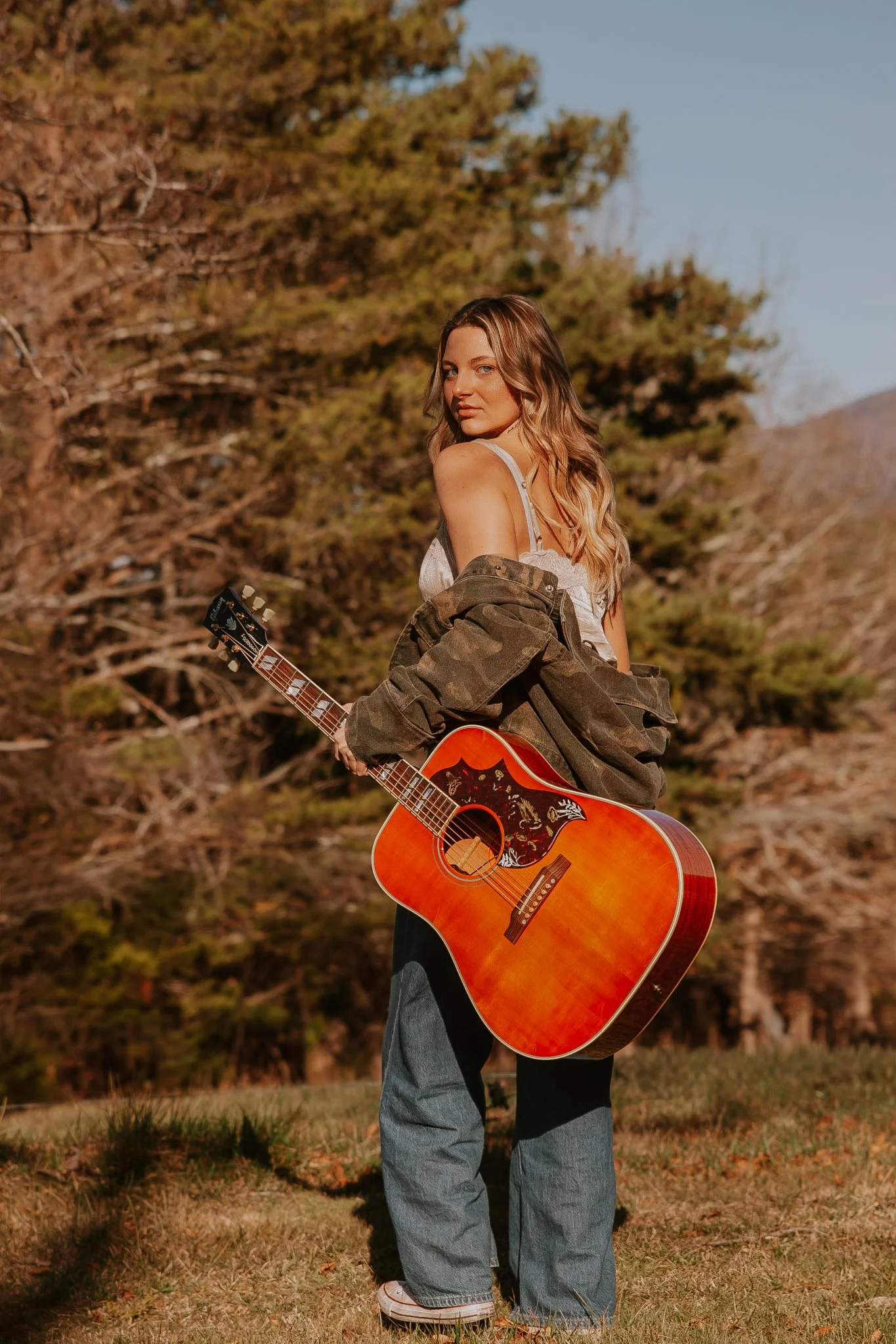 A young woman standing outdoors holding an acoustic guitar, wearing baggy jeans, a white tank top, and an oversized camouflage jacket draped over her shoulders, with trees and a mountain in the background.