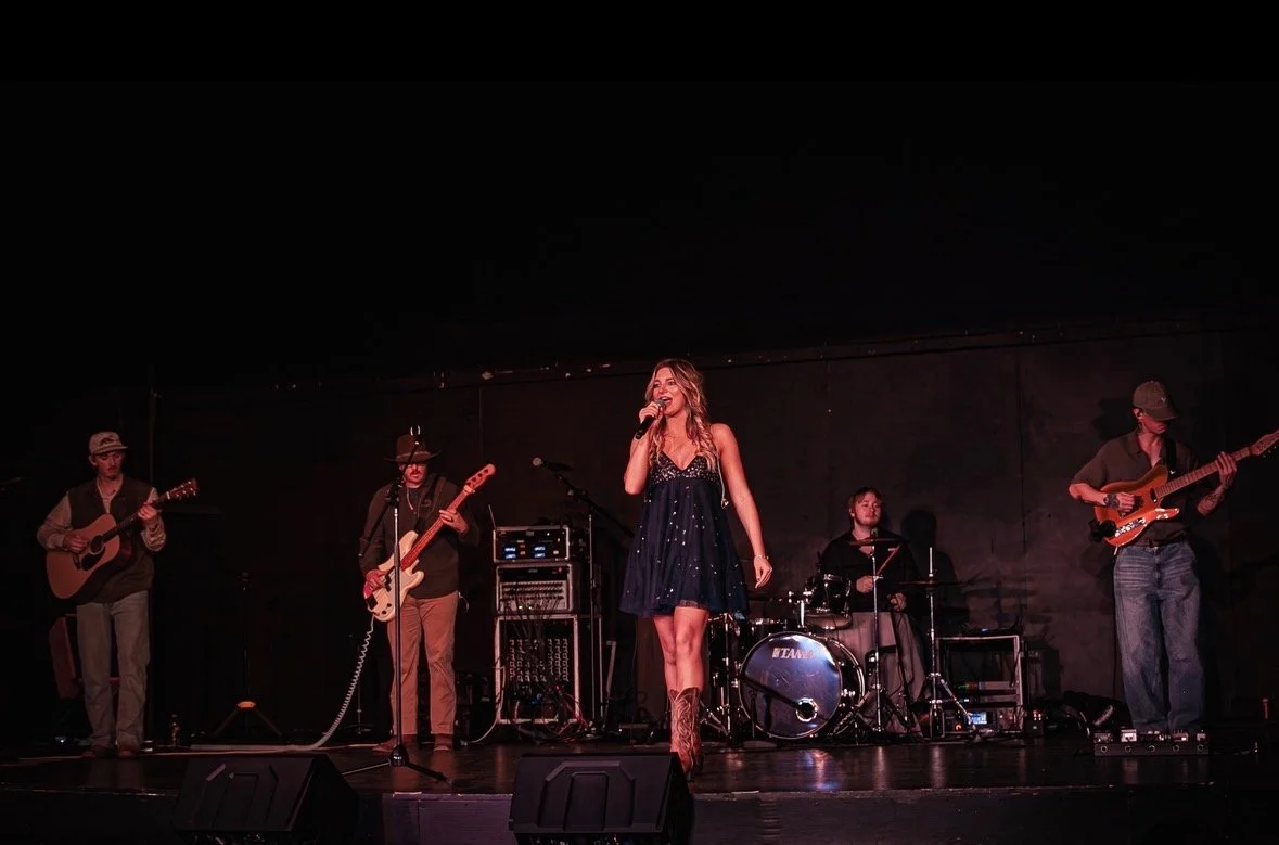 A woman singing on stage with a band, including two guitarists, a bassist, and a drummer.