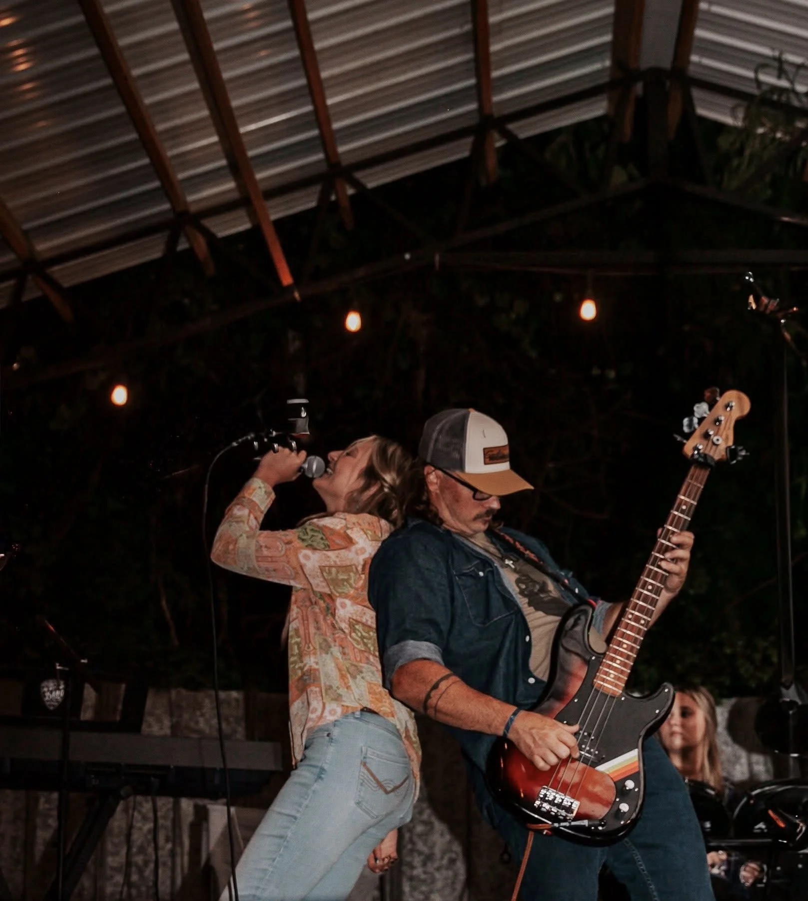 A woman singing into a microphone and a man playing an electric bass guitar on stage at a nighttime outdoor event. There are string lights and a keyboard in the background.