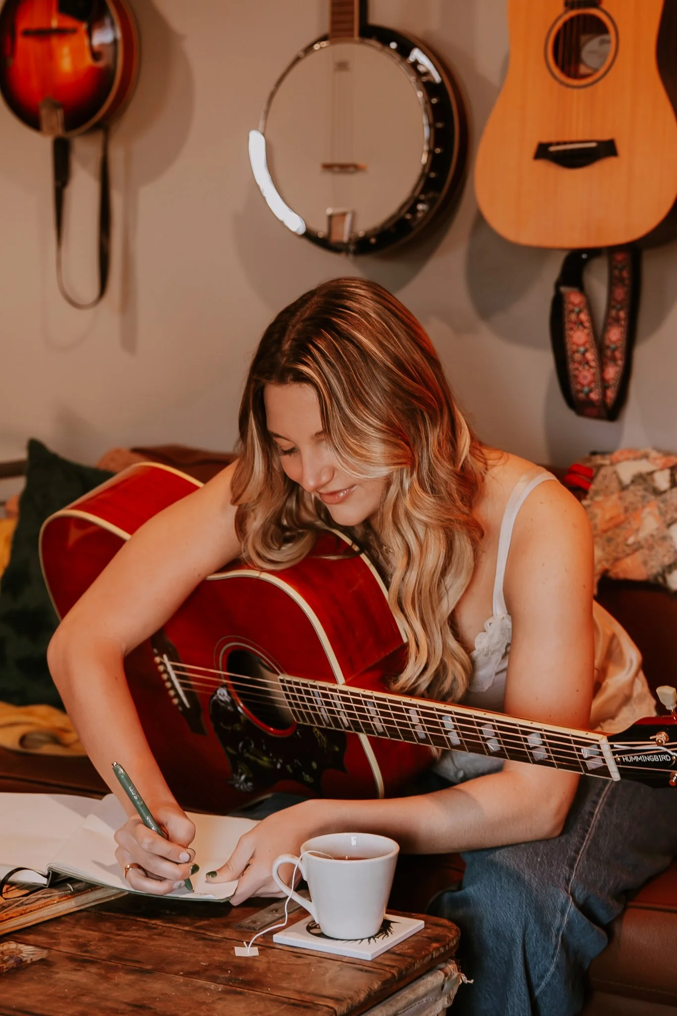 A woman with long wavy hair sitting on a couch, writing in a notebook, with a red acoustic guitar resting on her lap. There is a white mug on the table in front of her, along with a tea bag and a tea infuser.