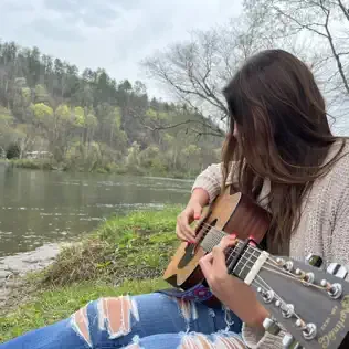 A woman sitting outdoors near a lake, playing an acoustic guitar surrounded by trees and greenery.