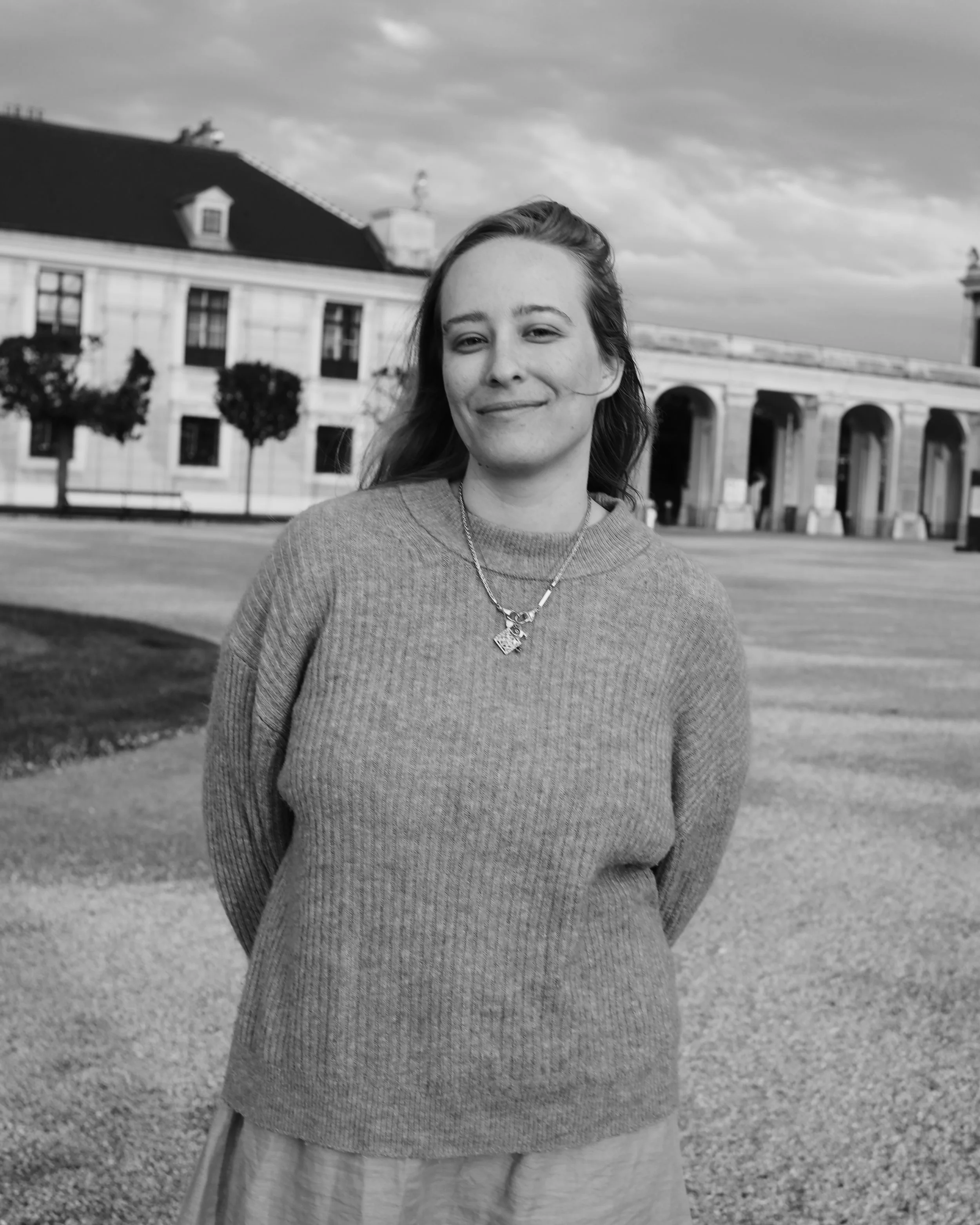 A woman smiling outdoors on a cloudy day with historic buildings and trees in the background.
