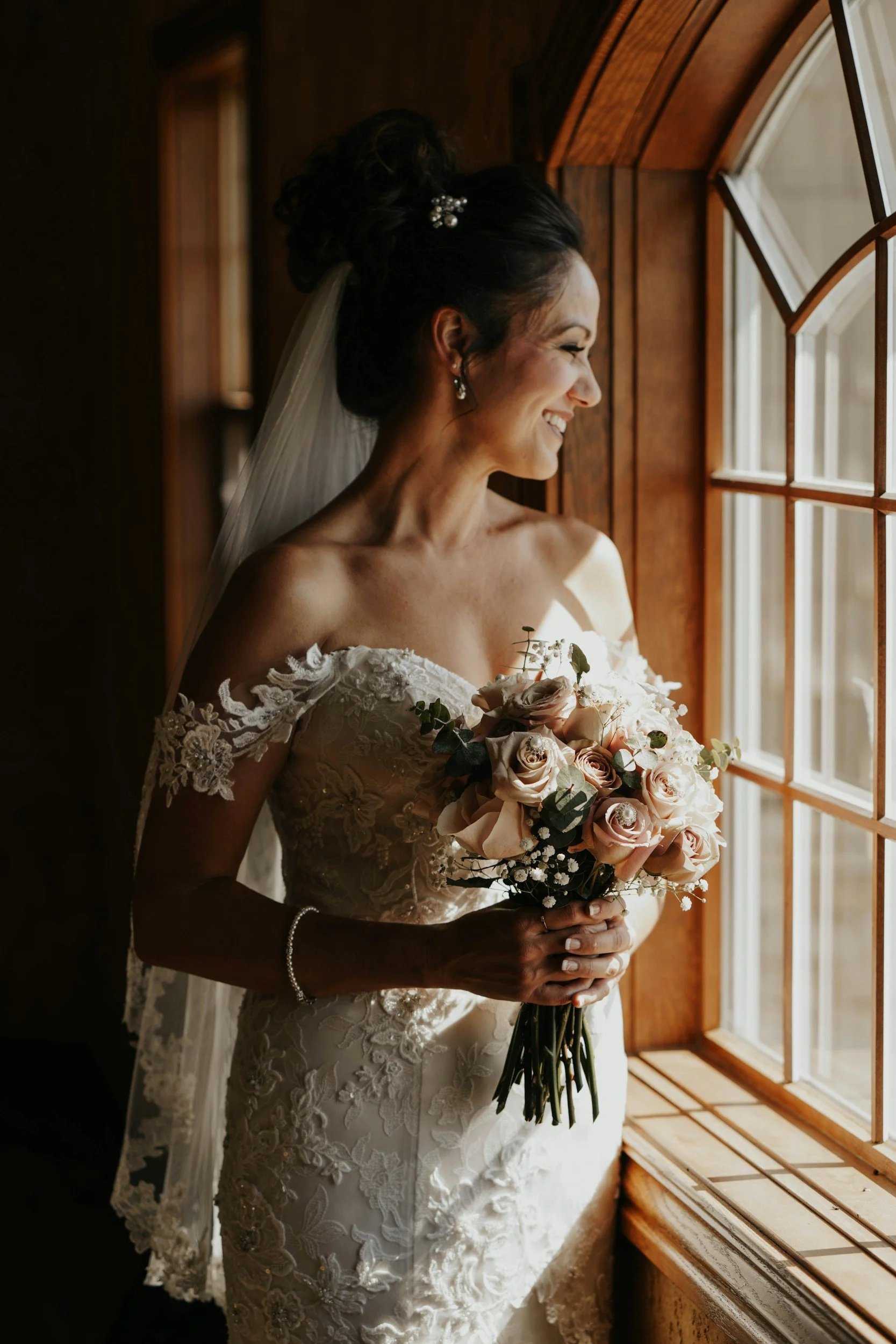 A smiling bride in a lace wedding dress holding a bouquet of pale pink roses and white flowers, standing by a window with wooden framing, looking outside with sunlight illuminating her profile.