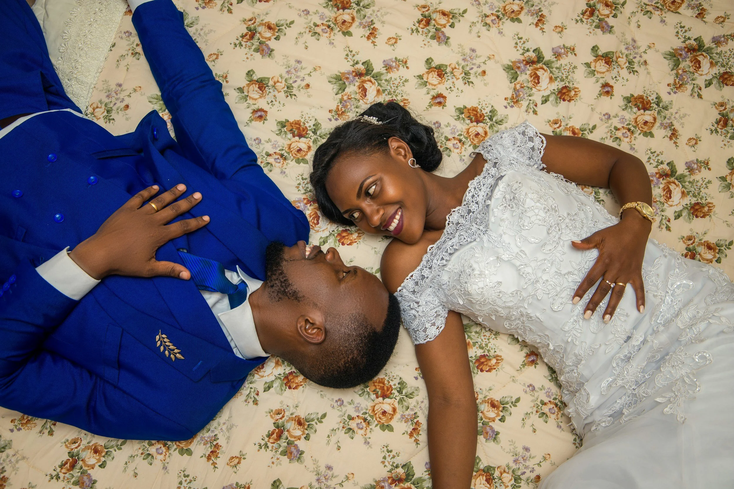 A couple dressed in formal attire lying on a floral bedspread, smiling and looking at each other.