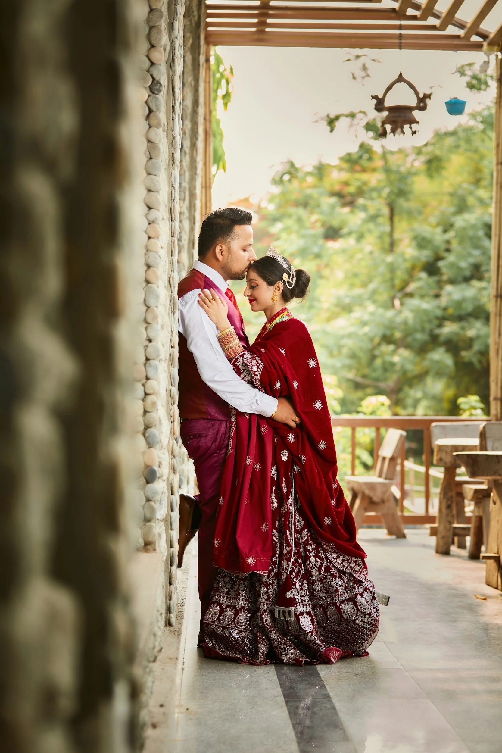 A couple dressed in traditional Indian wedding attire embracing in a serene outdoor setting with lush green trees in the background. The woman is wearing a red embroidered saree with gold jewelry and a tiara, and the man is in a maroon vest with a white shirt.