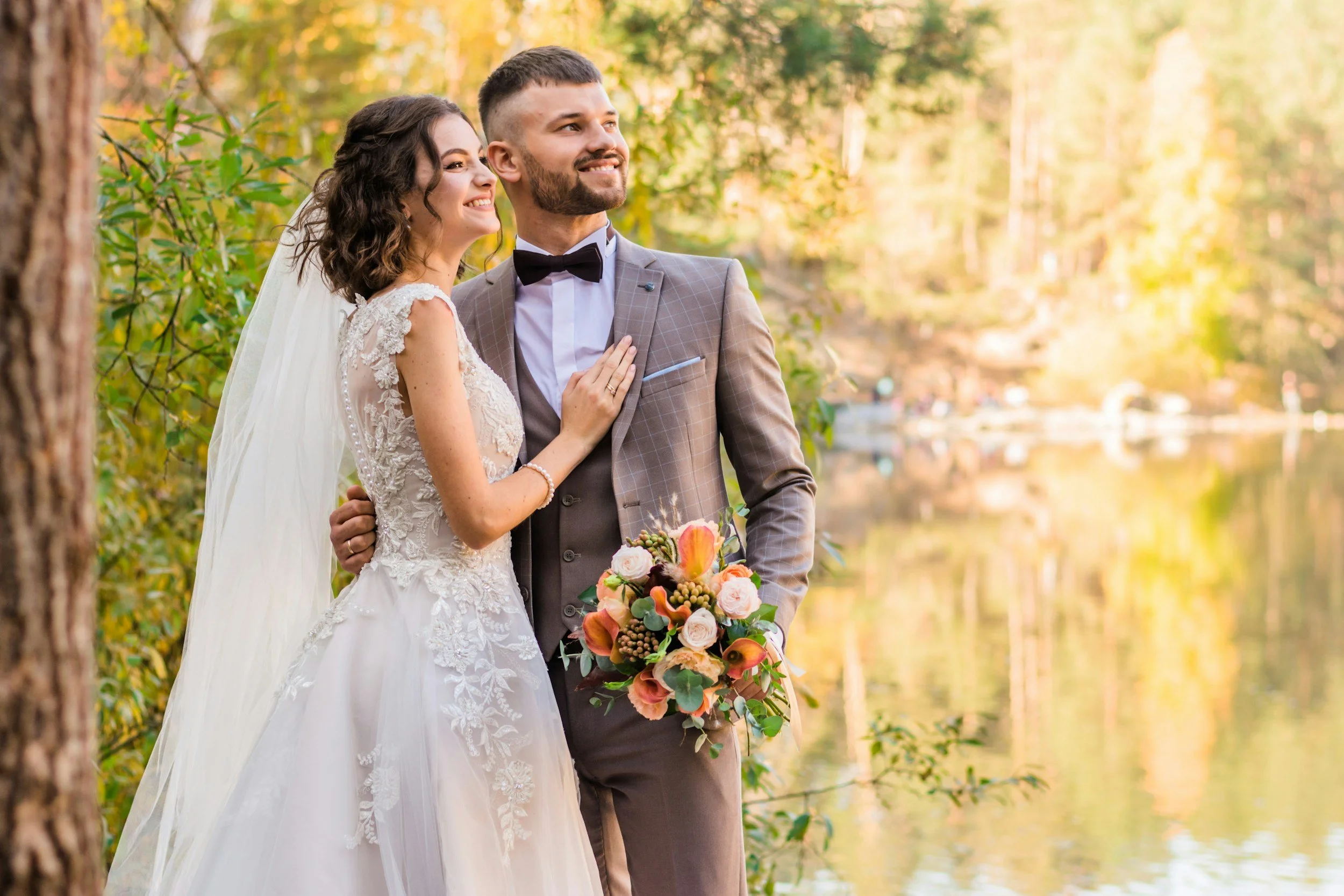A bride and groom standing close together outdoors in a forested area, smiling and looking happy. The bride has brown curly hair and is wearing a white wedding dress, while the groom has short dark hair, a beard, and is dressed in a brown checked suit with a black bow tie. The bride is holding a colorful bouquet of flowers.