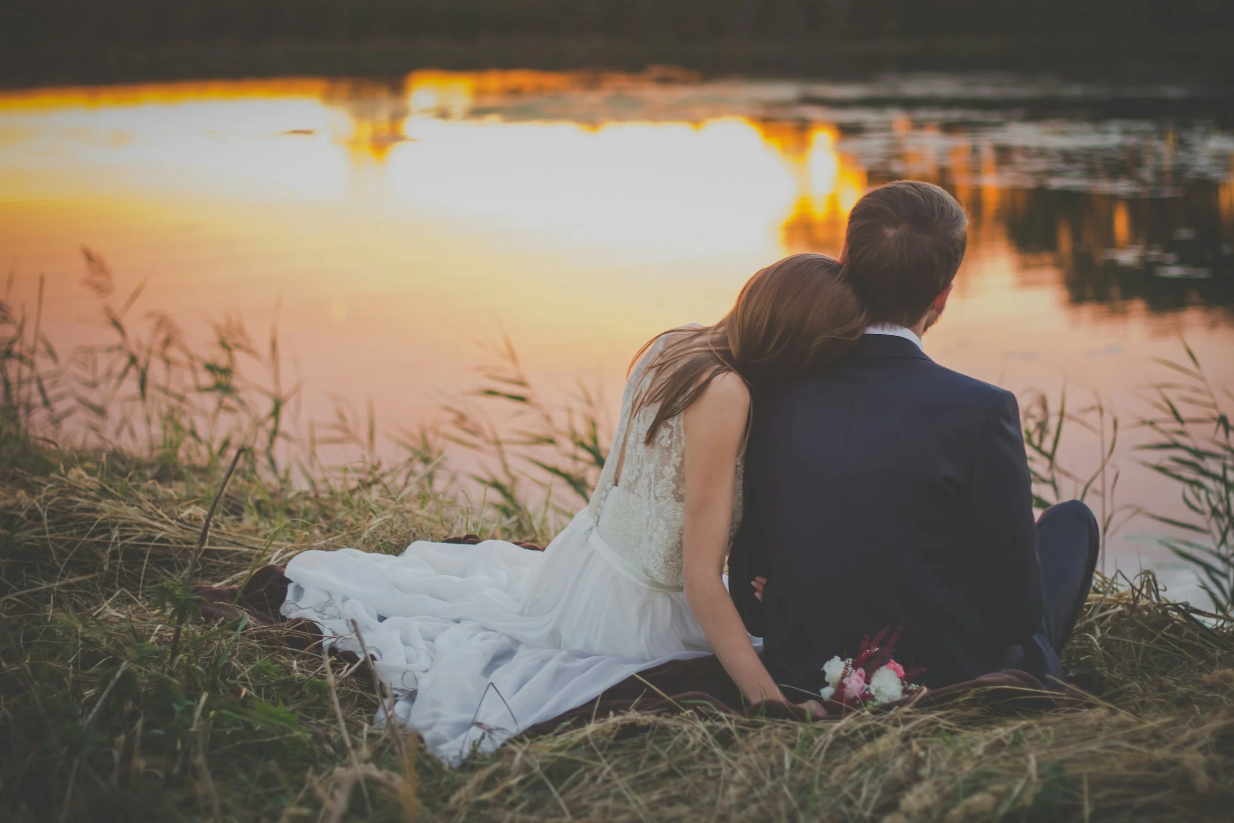 A bride and groom sitting by a lake at sunset, with the bride in a white wedding dress and the groom in a dark suit, embracing each other.