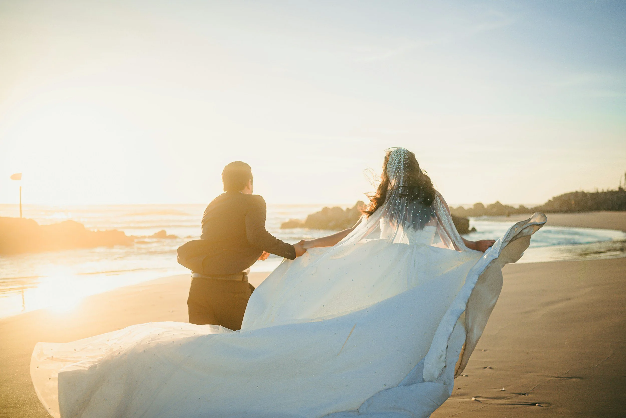 A couple sitting on the beach at sunset, holding hands, with a large white fabric or dress flowing around them.