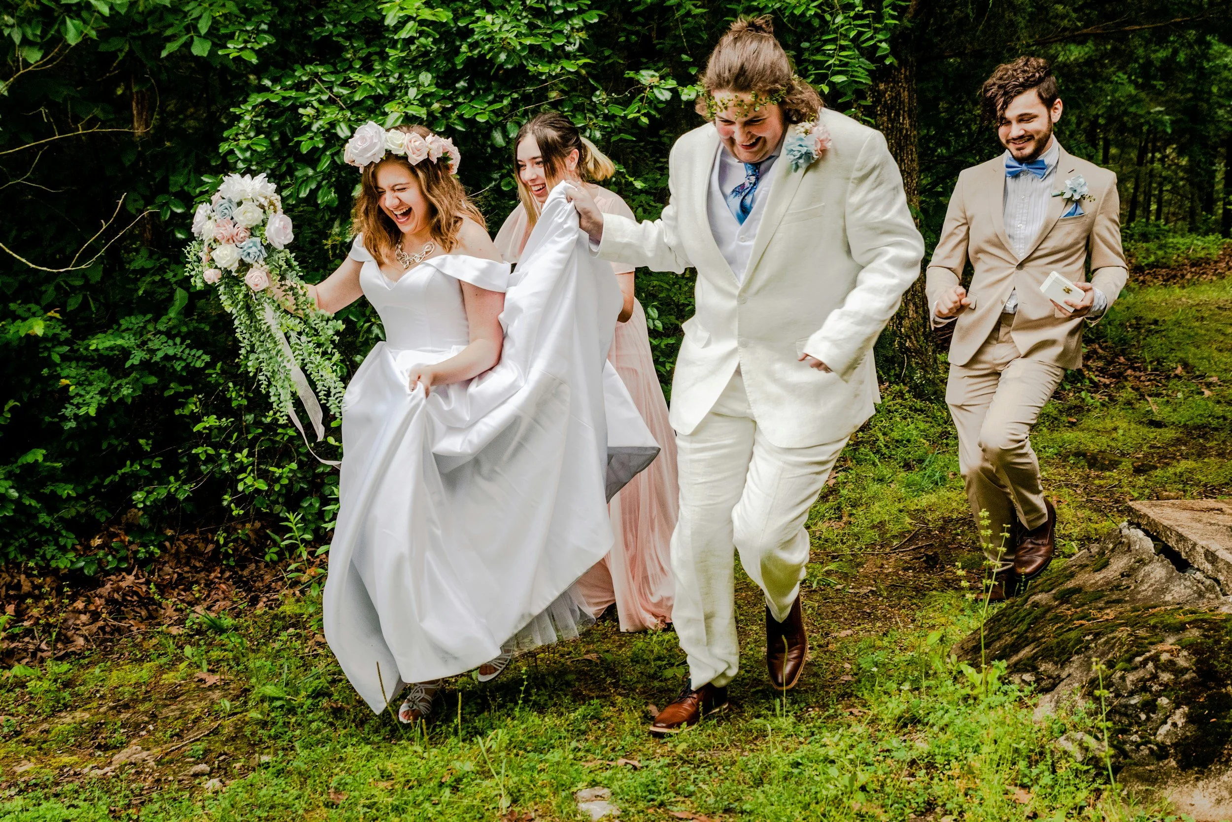 Group of five people in wedding attire walking outdoors through a wooded area, smiling and laughing.