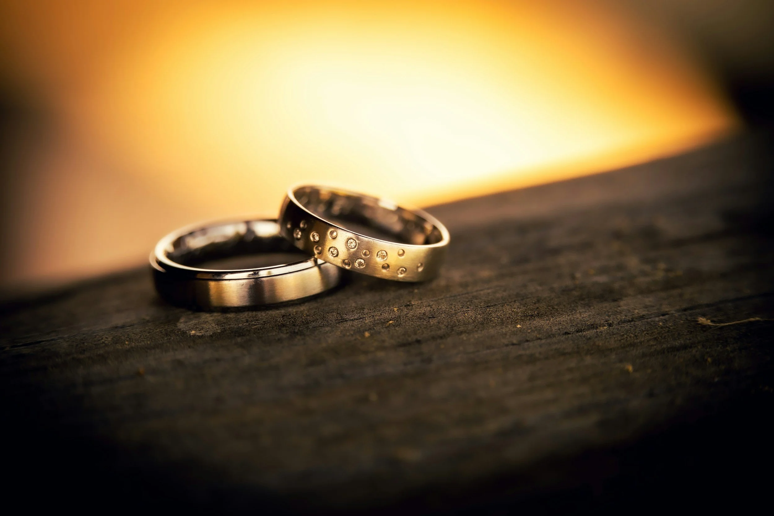 Close-up of two wedding rings resting on a dark wooden surface with a blurred warm light background.