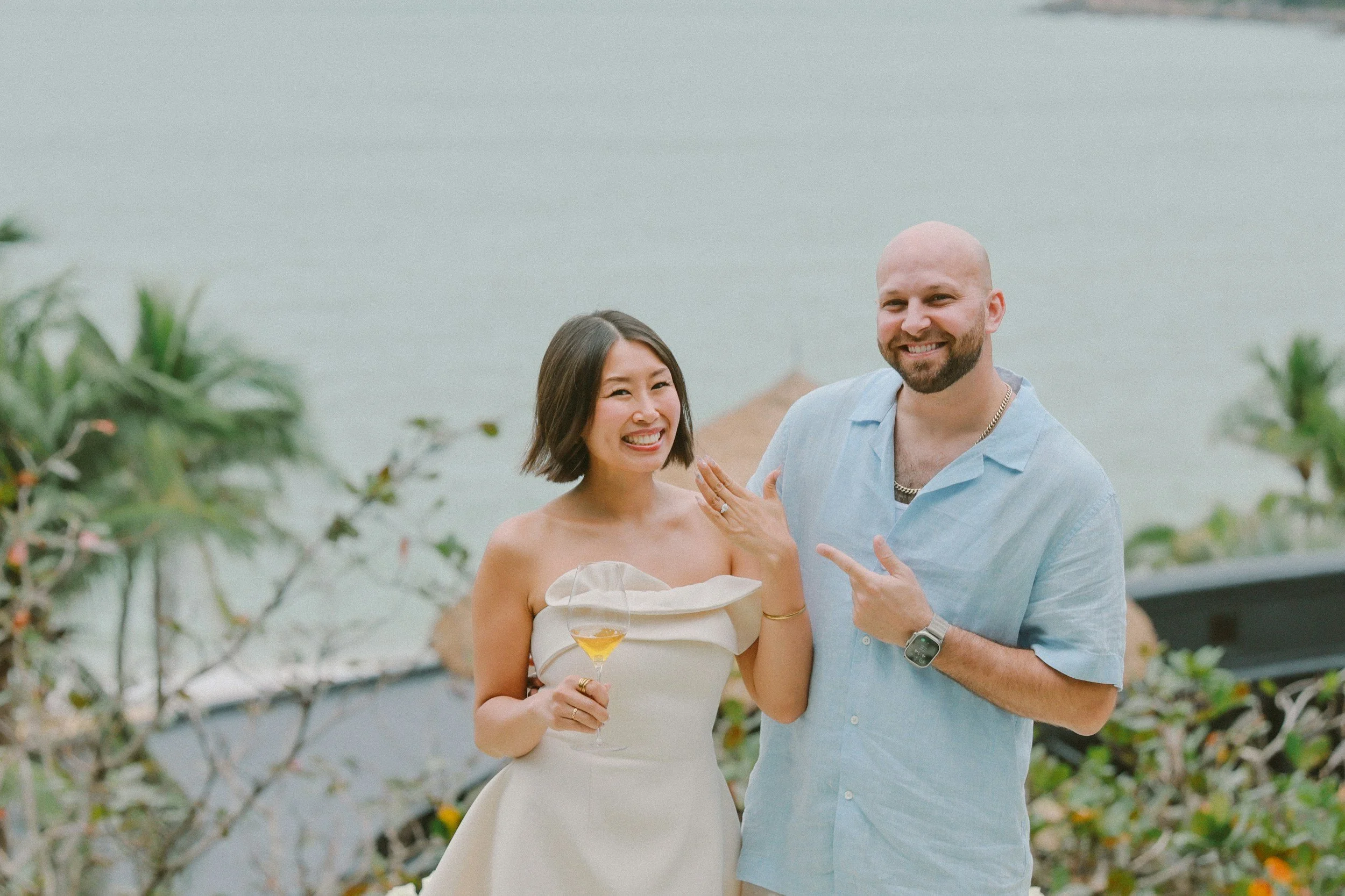 A smiling woman in a strapless white dress holding a drink, standing next to a smiling man with a beard in a light blue shirt, outdoors near a body of water and greenery.