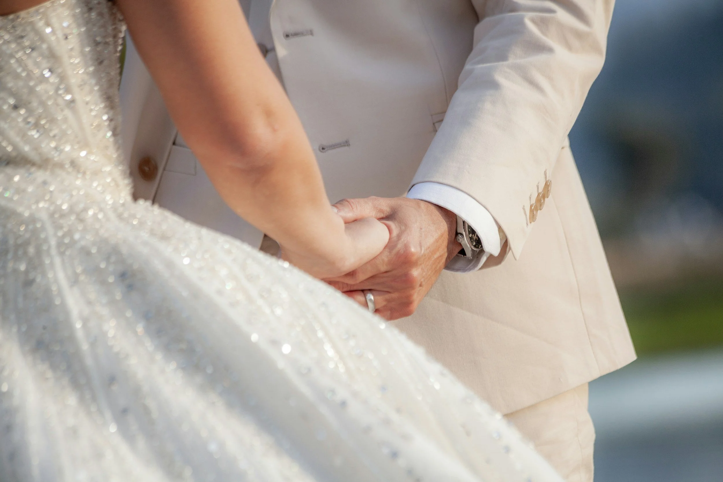 Close-up of a wedding couple holding hands, with the bride wearing a sparkly white dress and the groom dressed in a beige suit and white shirt.