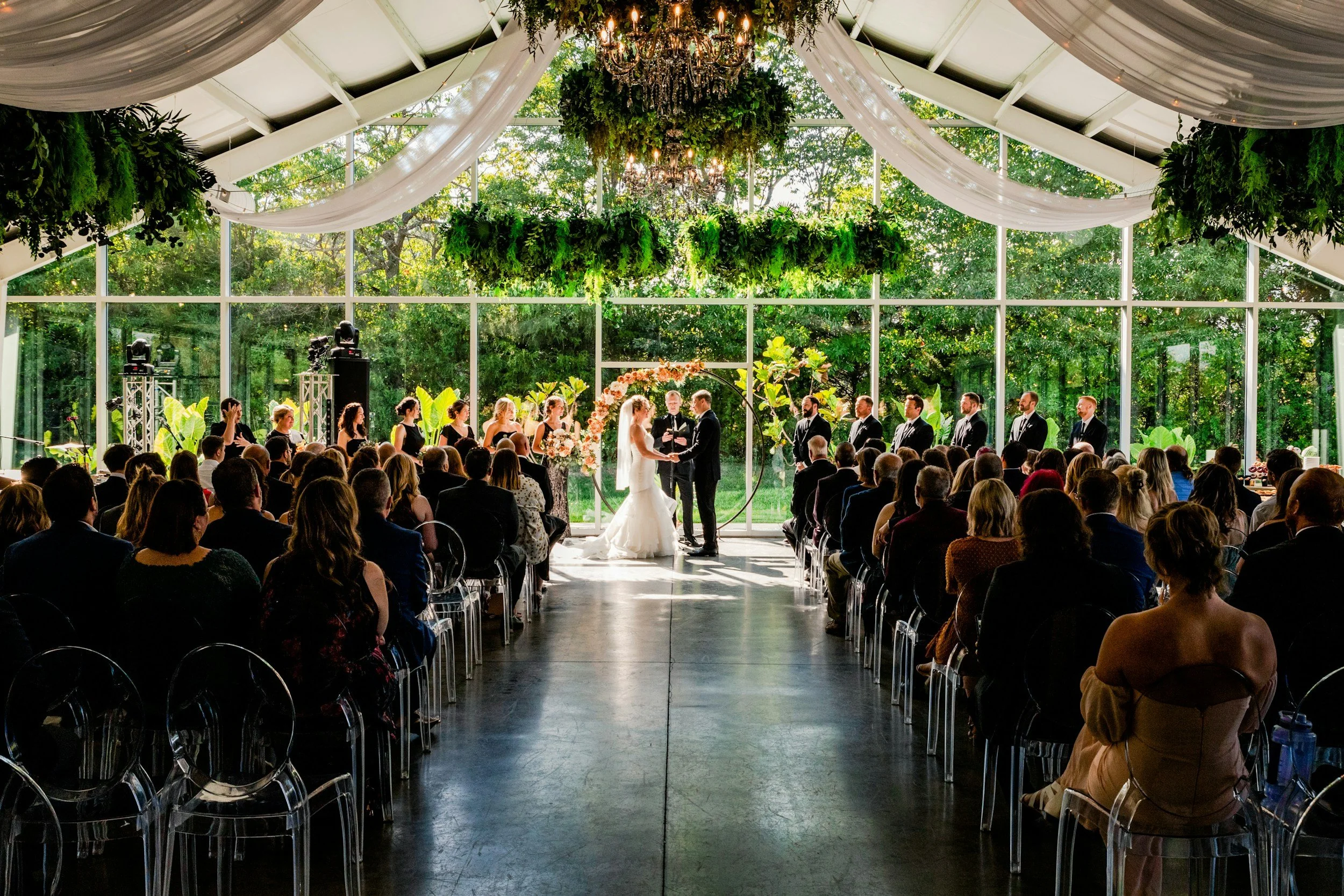 Indoor wedding ceremony with a bride and groom standing under a floral arch, surrounded by bridesmaids and groomsmen, with guests seated and watching.