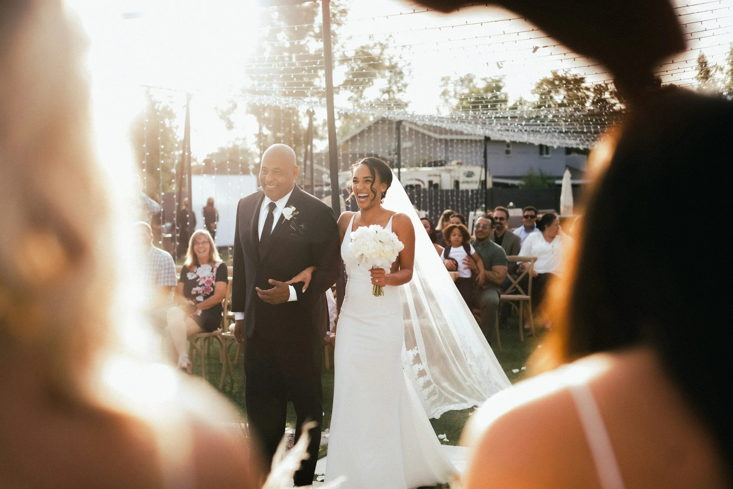 A bride and groom walking down the aisle at their outdoor wedding ceremony, smiling, with guests seated in the background, decorated with string lights on a sunset evening.