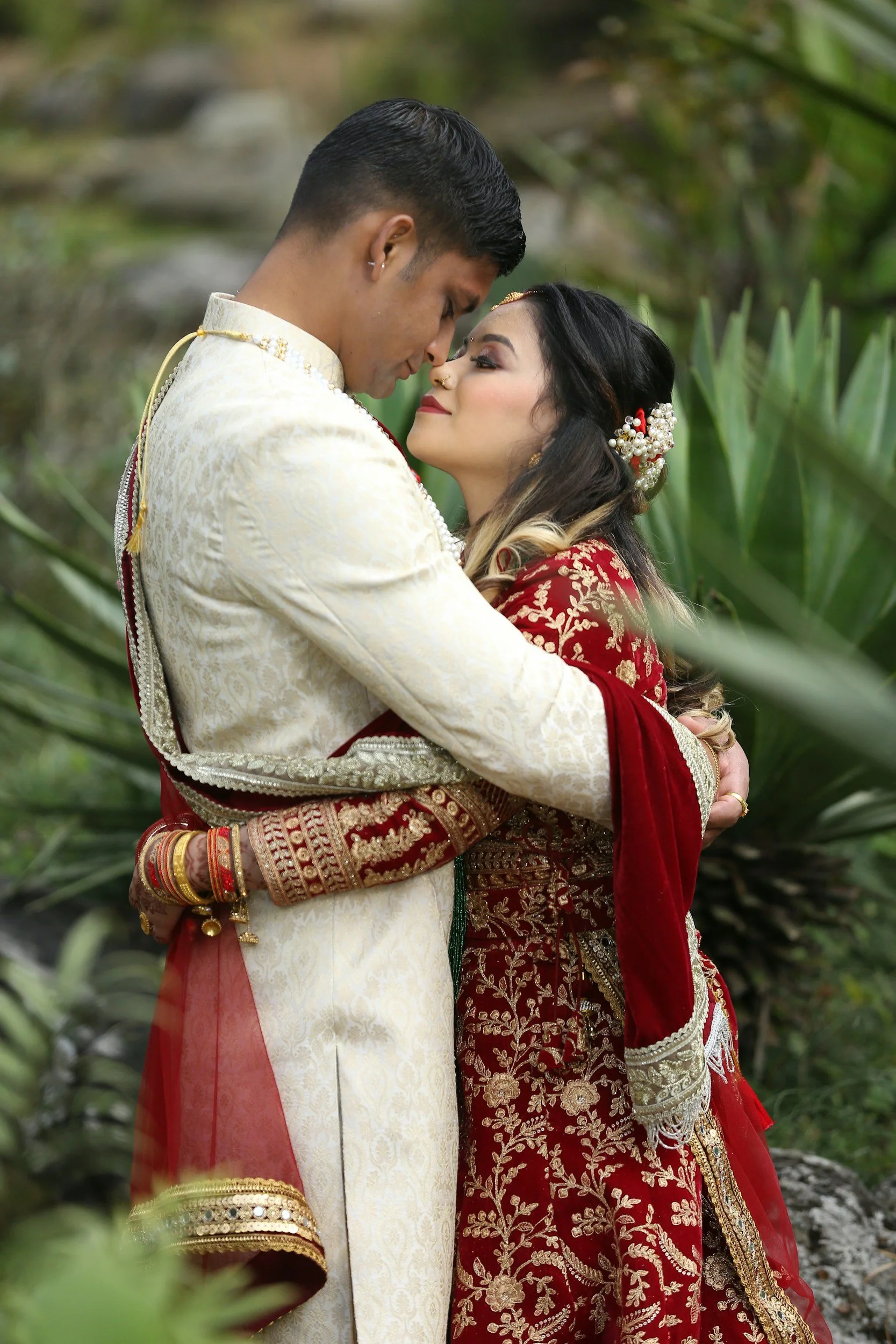 A couple dressed in traditional South Asian wedding attire embracing outdoors surrounded by greenery.