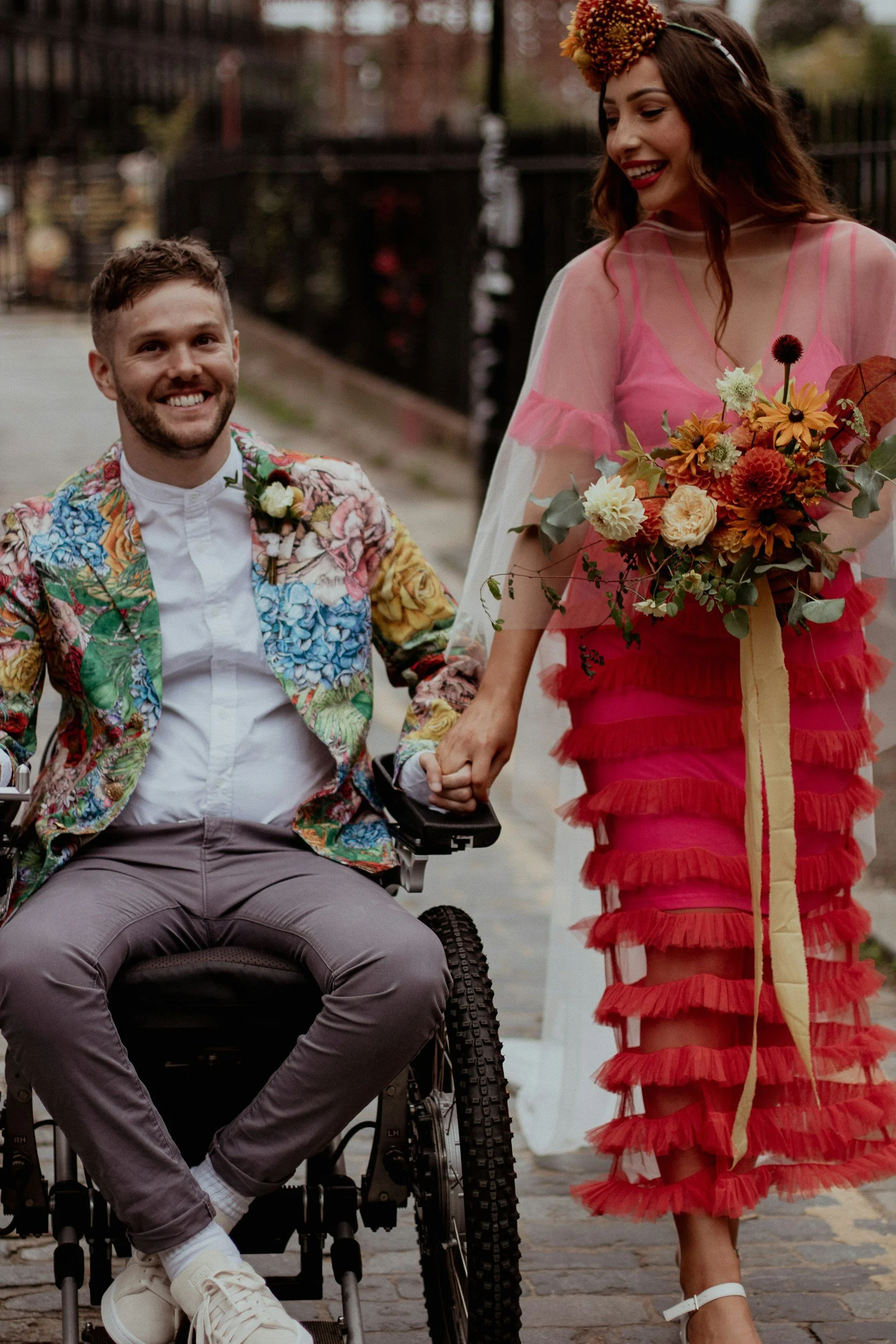 A smiling man in a wheelchair holding hands with a smiling woman. The man wears a colorful floral blazer and a white shirt, while the woman wears a sheer pink top with ruffles and a vibrant red layered skirt, holding a bouquet of flowers and wearing a floral headpiece. They are outdoors in a city setting with a fence and trees in the background.
