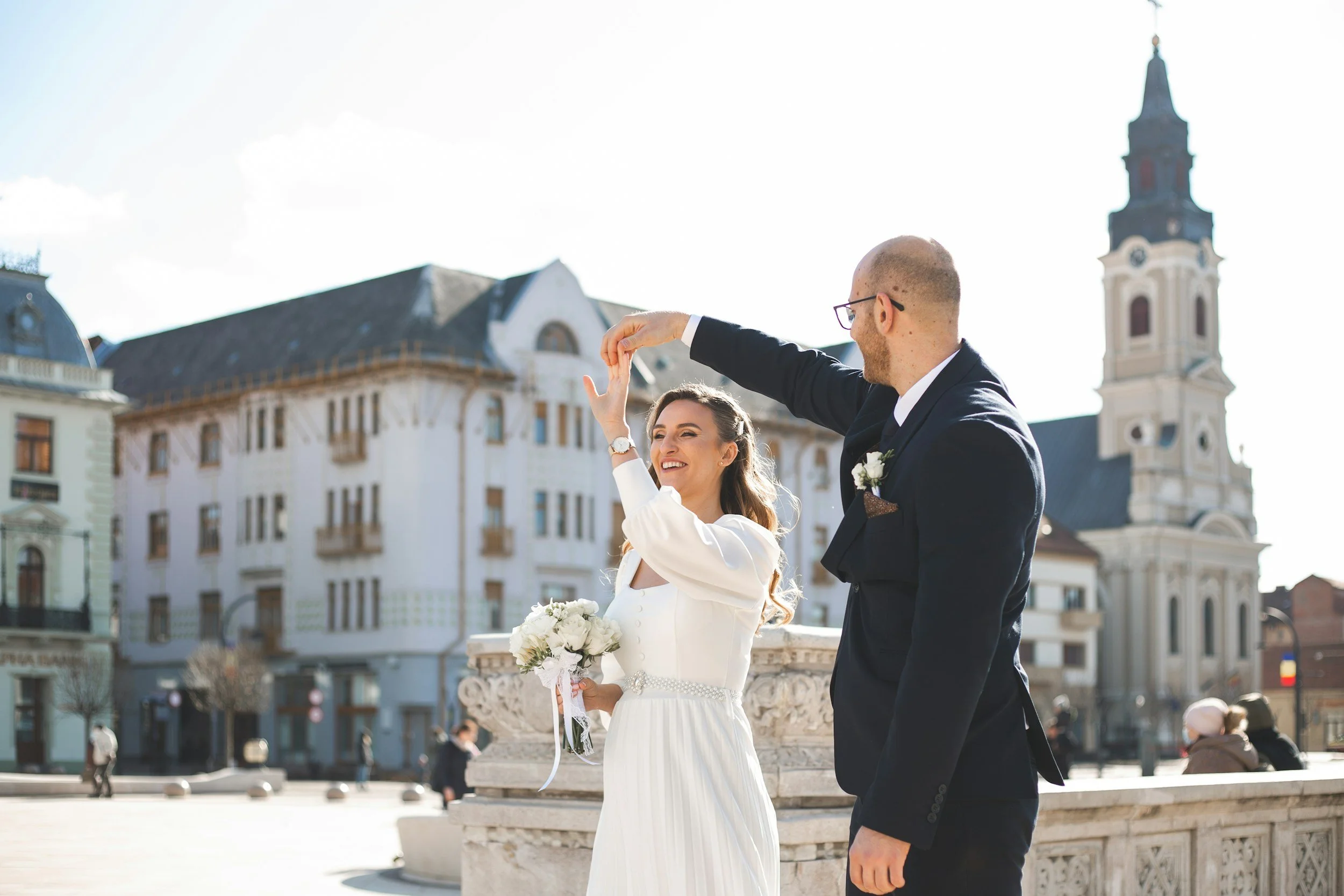 A bride and groom dancing outdoors in a European city square with historic buildings and a church tower in the background.