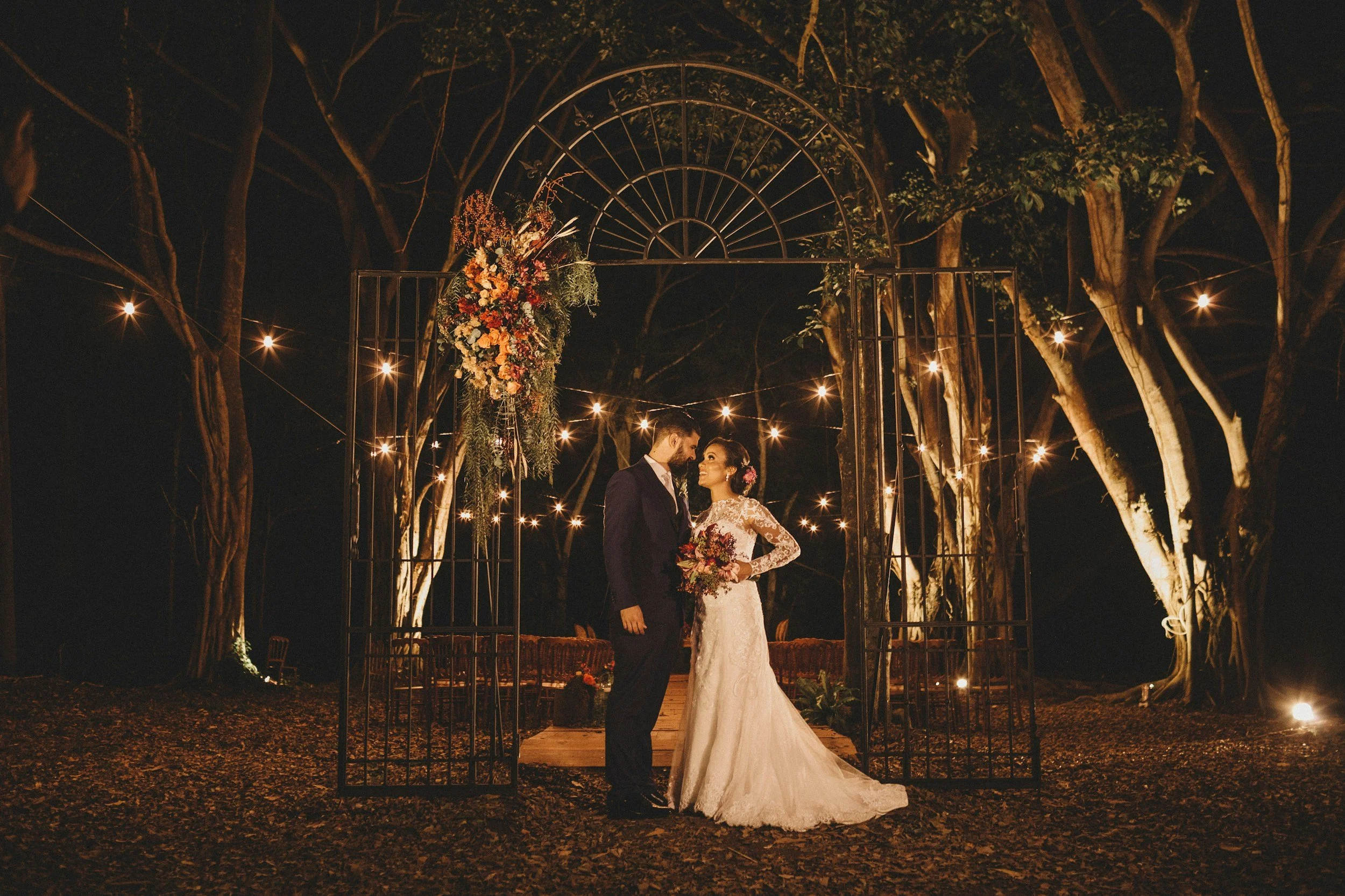 A bride and groom stand together under a decorative metal arch at night, surrounded by string lights and trees, during their outdoor wedding ceremony.