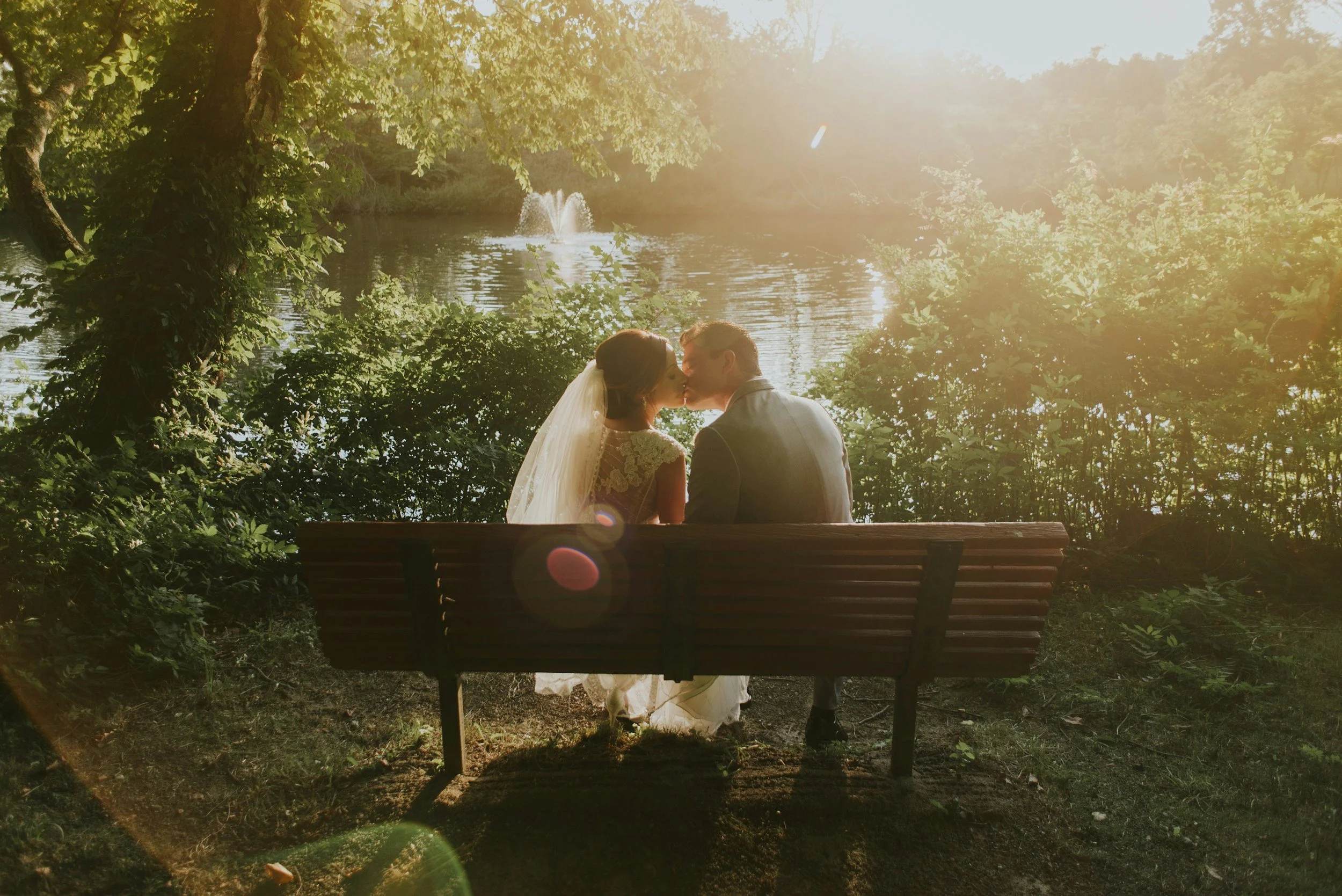 A couple is sitting on a park bench kissing, with a lake and fountain in the background, surrounded by trees and sunlight.