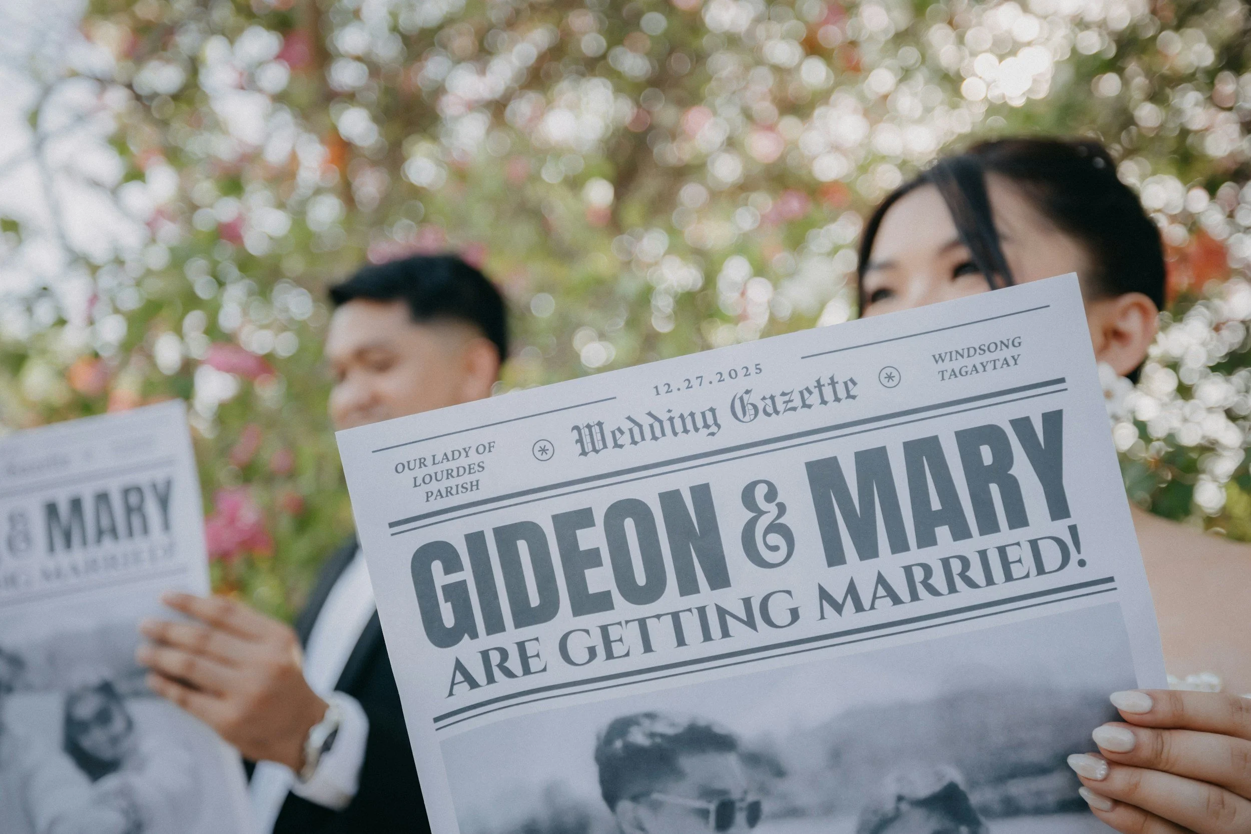 A woman with black hair holding a wedding announcement sign at an outdoor wedding. There are blurry trees with pink flowers in the background.