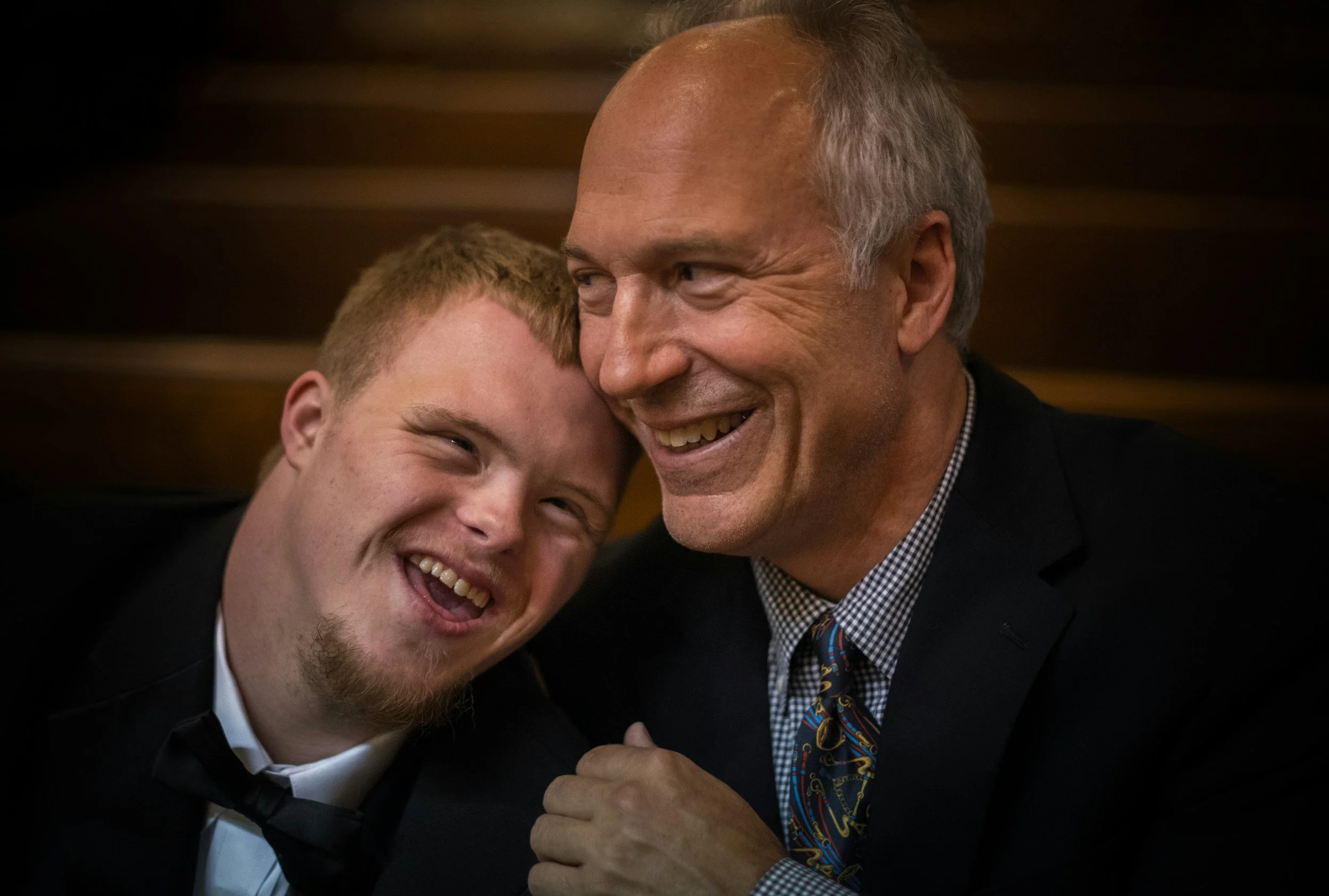 An older man with gray hair and a young man with red hair are smiling and leaning their heads together in a warm embrace, showcasing a moment of joy and friendship.