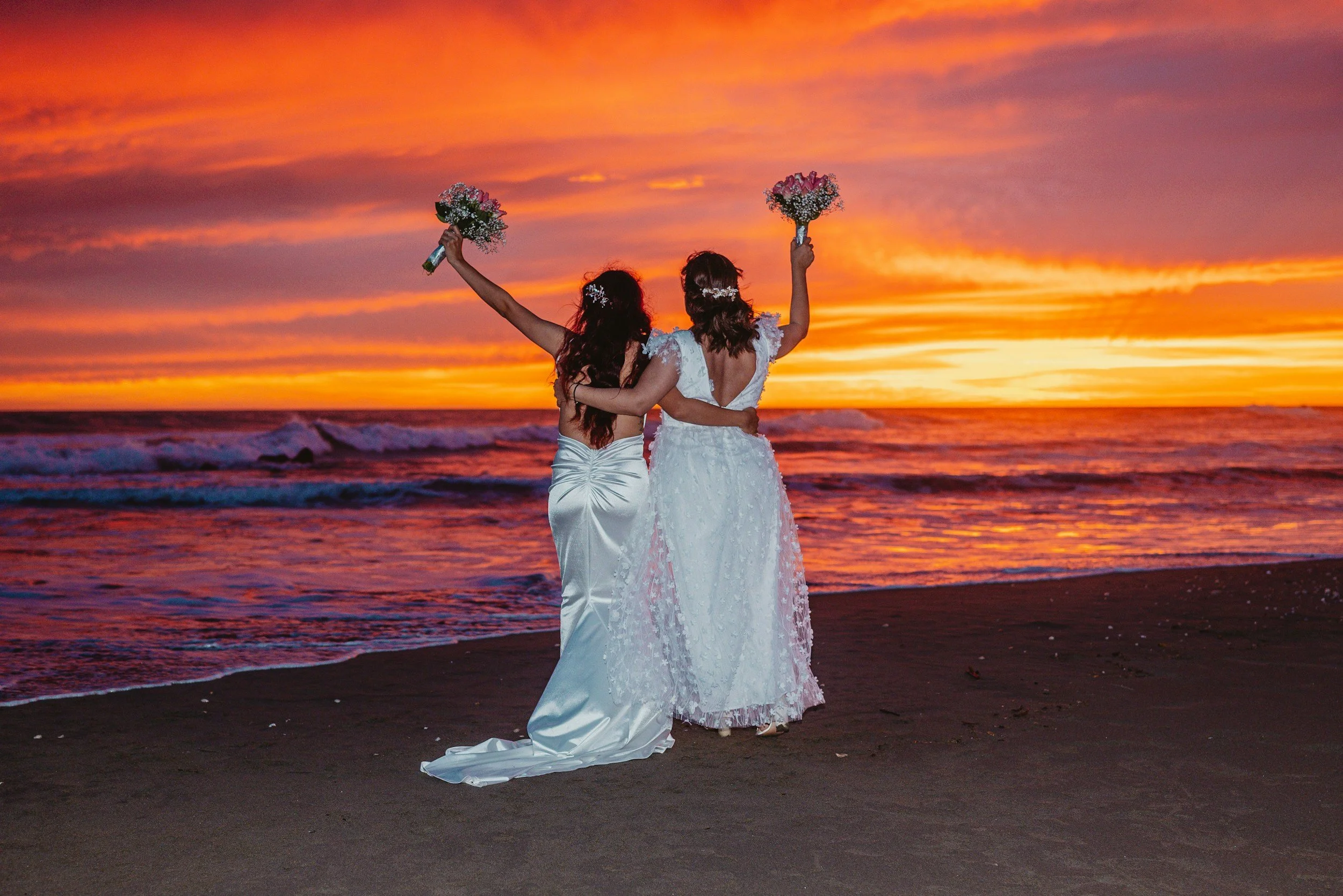 Two women in wedding dresses standing on a beach during sunset, holding bouquets, one with her arm around the other.