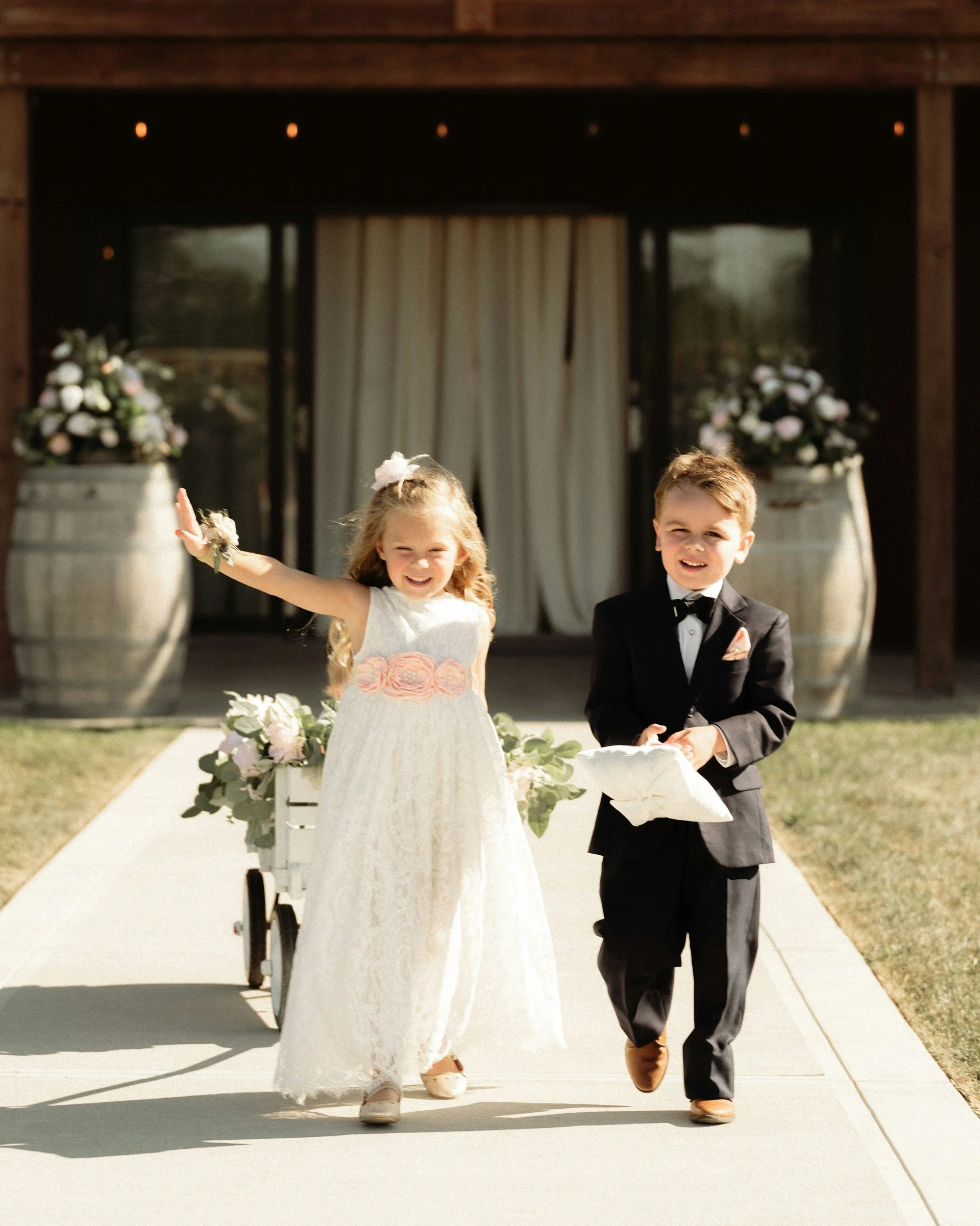 Two children, a girl in a white dress with pink flower accents and a boy in a tuxedo, are joyfully walking outdoors during a wedding ceremony. The girl is smiling and raising her hand, while the boy is smiling and holding a small item in his hand. Behind them are large white planters with flowers and a wedding backdrop with curtains.