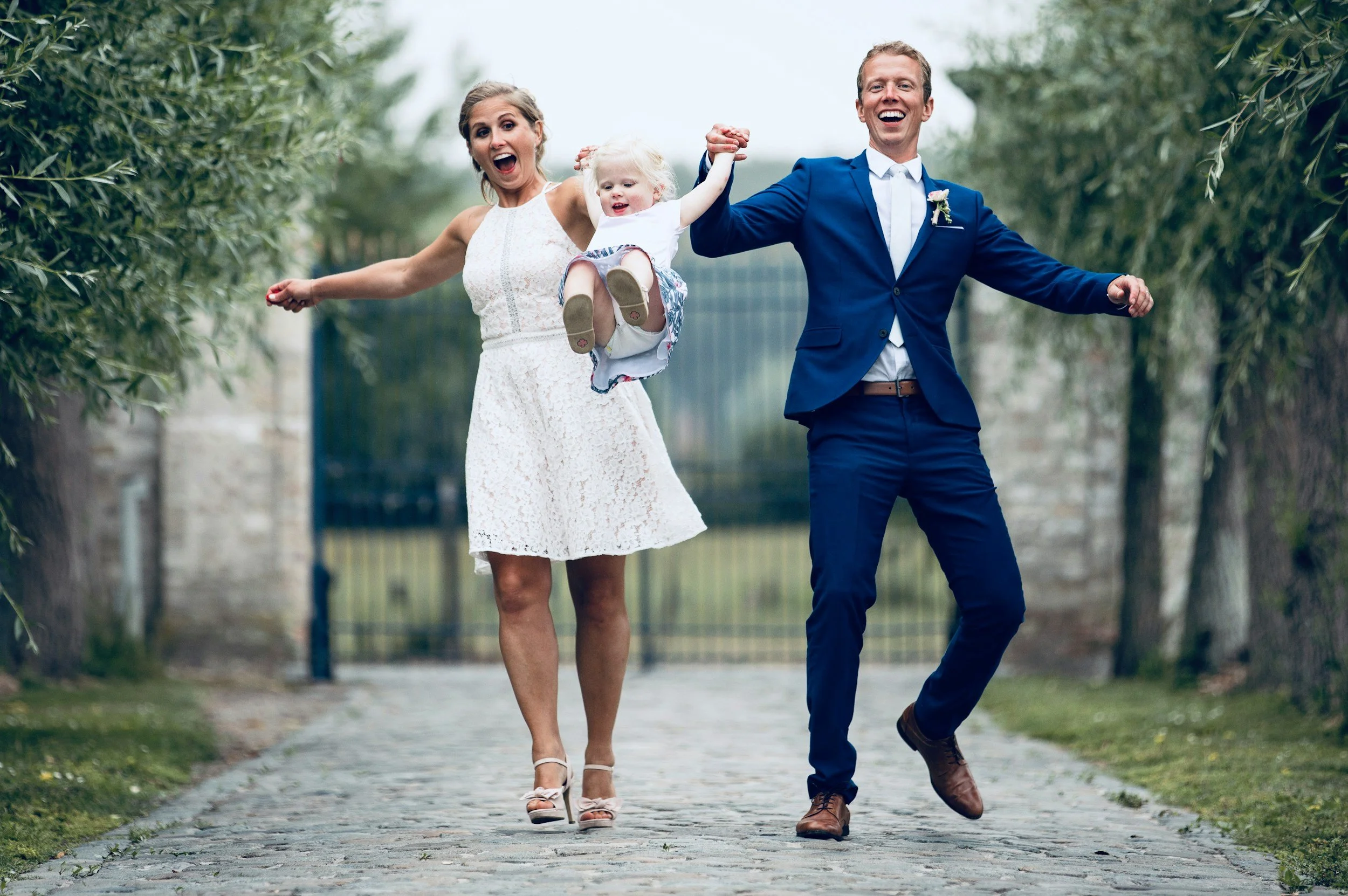 A happily married couple, a woman in a white lace dress and a man in a blue suit, holding hands and a young girl with blonde hair, wearing a white dress, walking down a cobblestone path surrounded by greenery and trees.