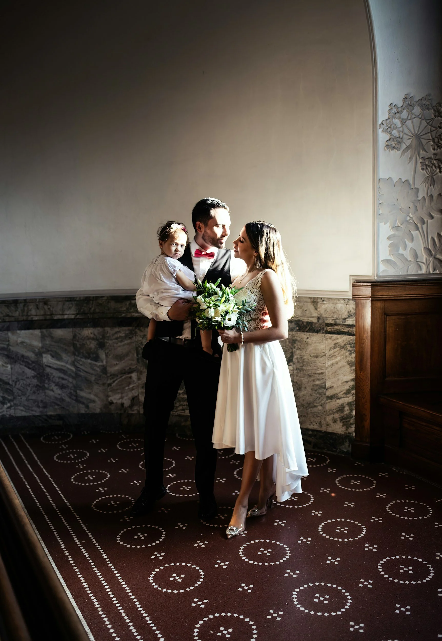 A bride and groom standing together with a young girl at a wedding, the groom holding a bouquet of flowers, in a room with marble lower walls and patterned carpet.