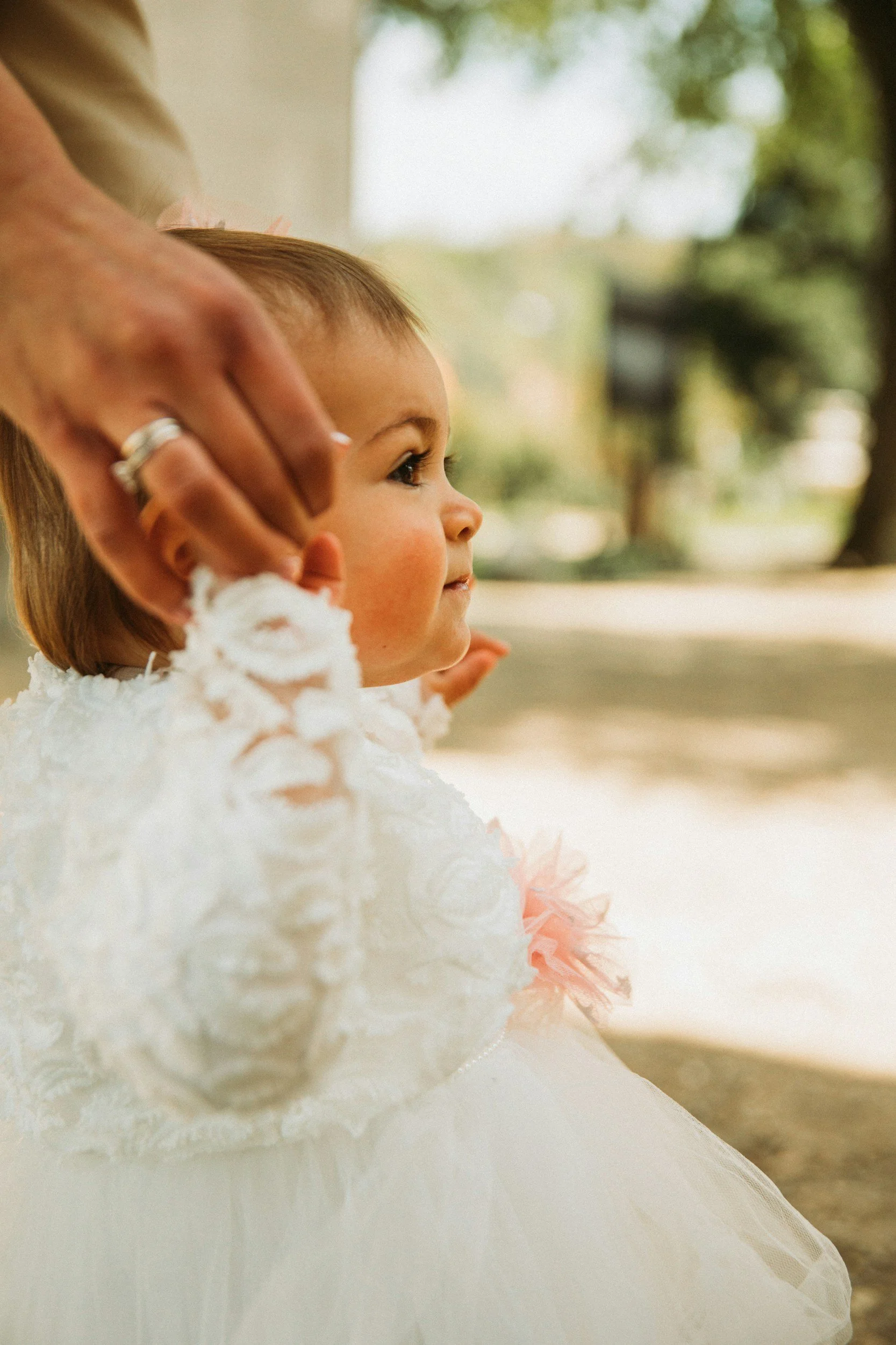 A young girl with light brown hair and a fair complexion, wearing a white dress with pink accents, sitting outdoors with a blurred background of trees and greenery.