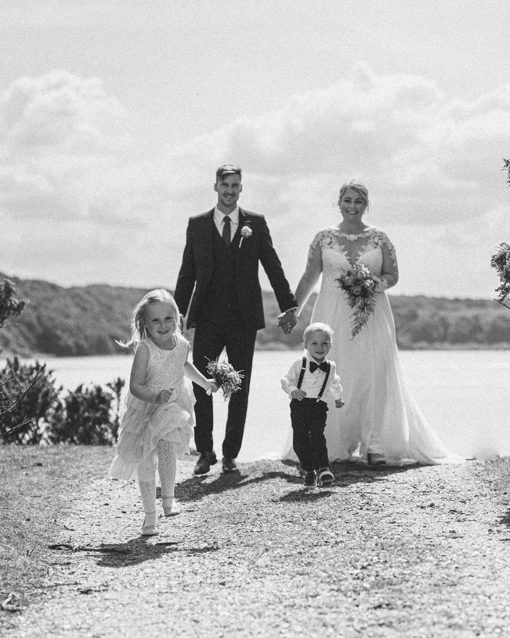 A bride and groom holding hands, walking outdoors on a gravel path with three children, a girl in a white dress and two boys in formal attire, under a partly cloudy sky.