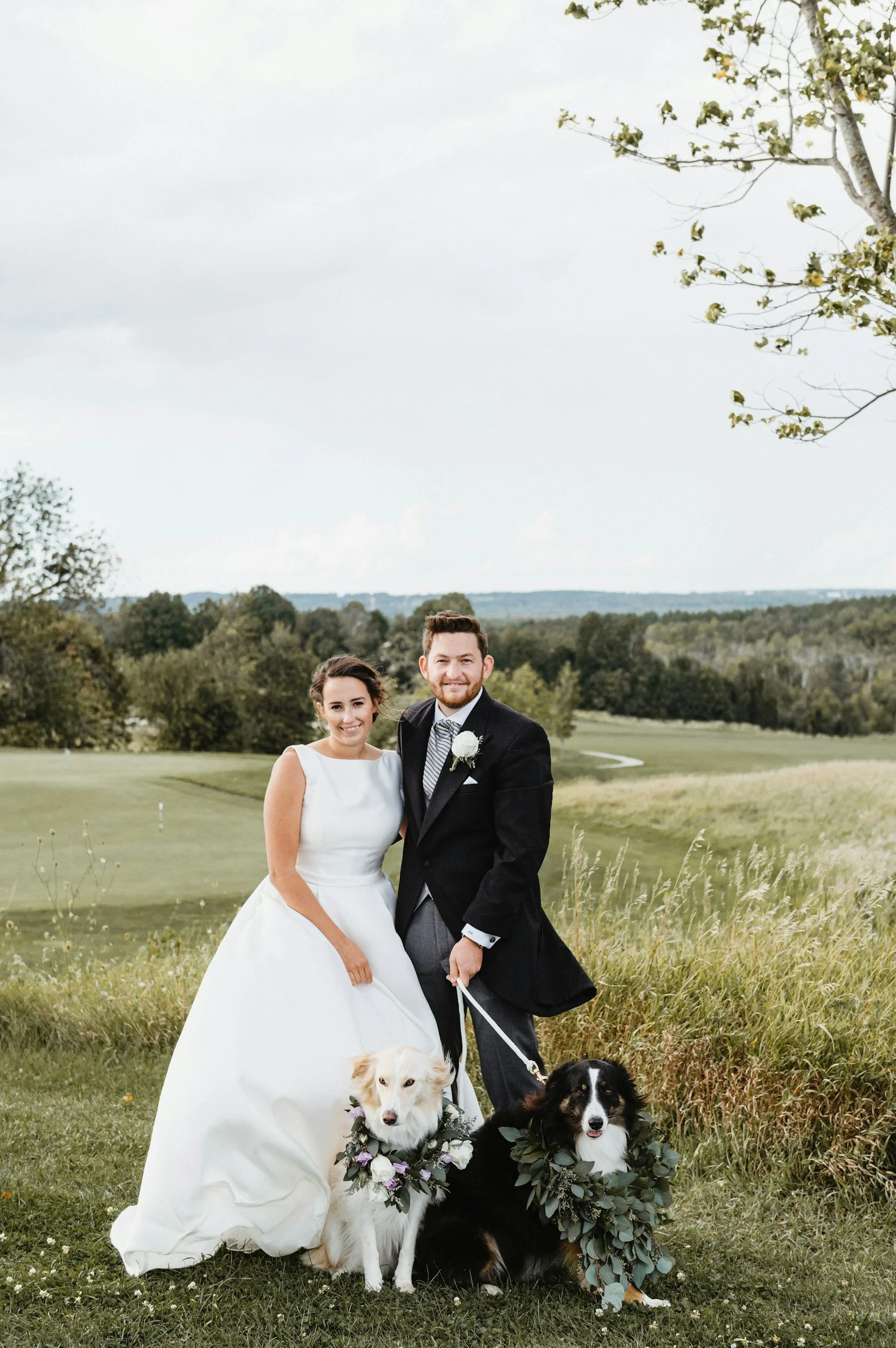 A bride and groom standing outdoors on a grassy field, with two dogs wearing wreaths of greenery and flowers, during a wedding photoshoot, with a backdrop of trees and hills under an overcast sky.