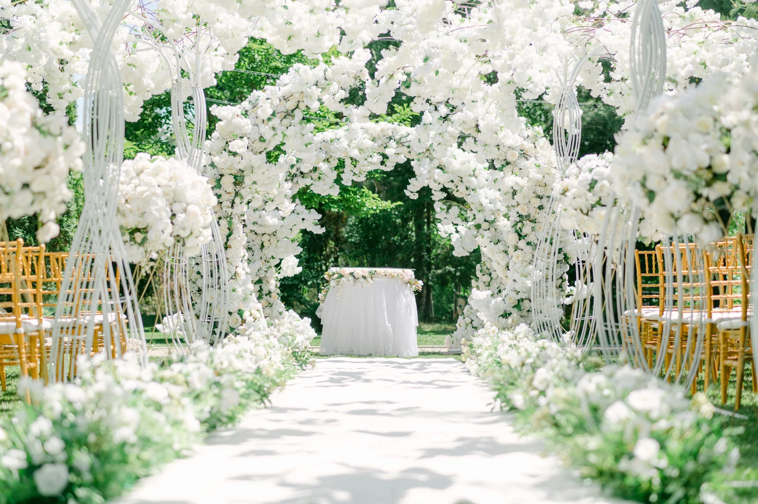 A wedding aisle decorated with white flowers on both sides, leading to an altar decorated with flowers, set outdoors with trees and green foliage in the background.