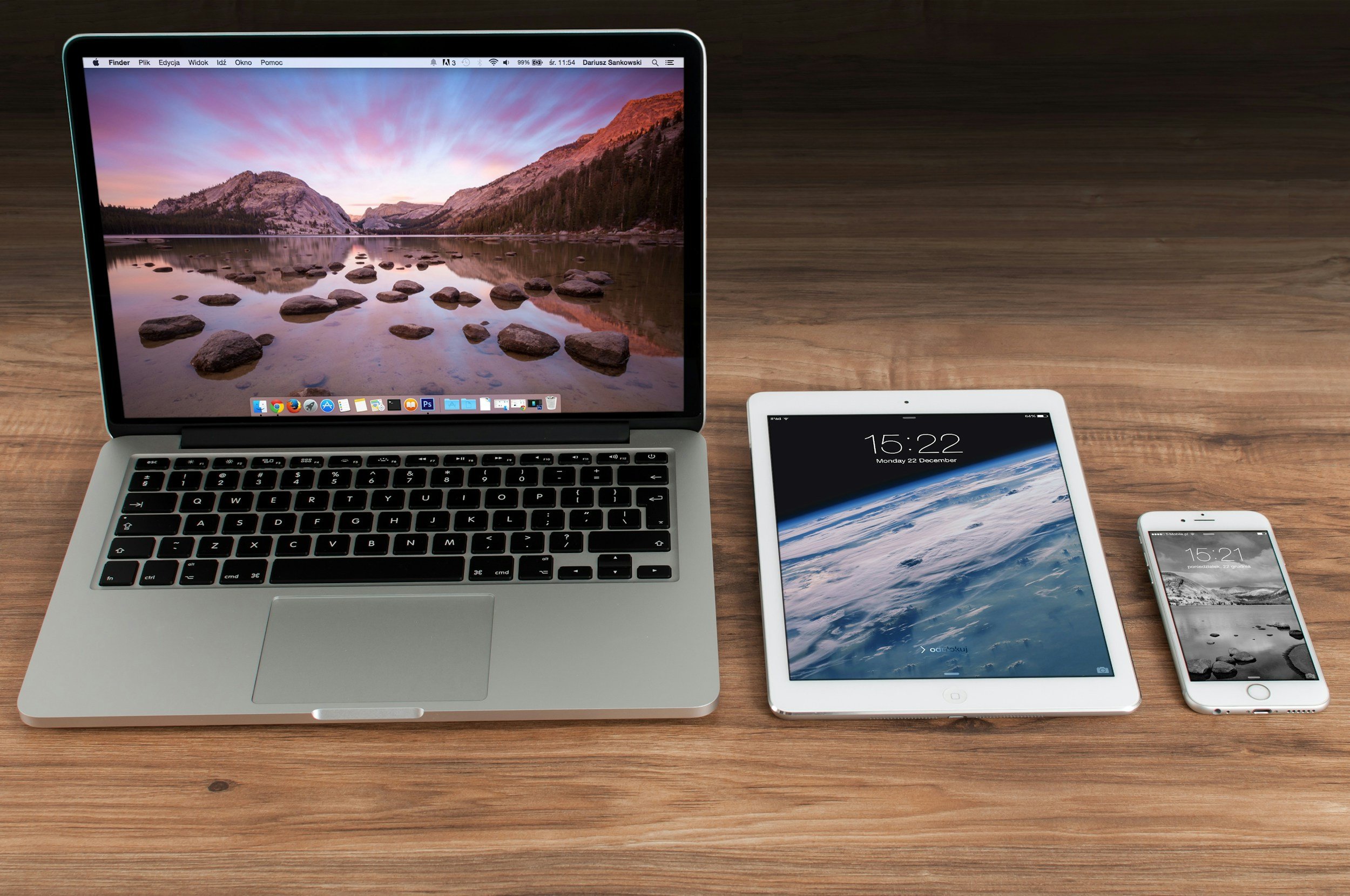 An Apple MacBook laptop, an iPad, and an iPhone on a wooden table, all displaying scenic wallpapers of mountains and skies.