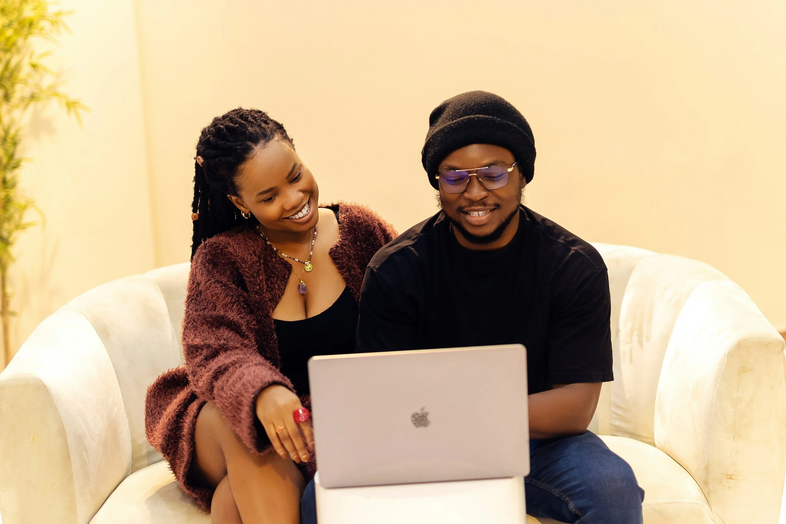 A woman with styled hair and jewelry, sitting on a cream sofa, taking a selfie with a young man wearing glasses and a beanie, using a silver laptop in a cozy, well-lit room.