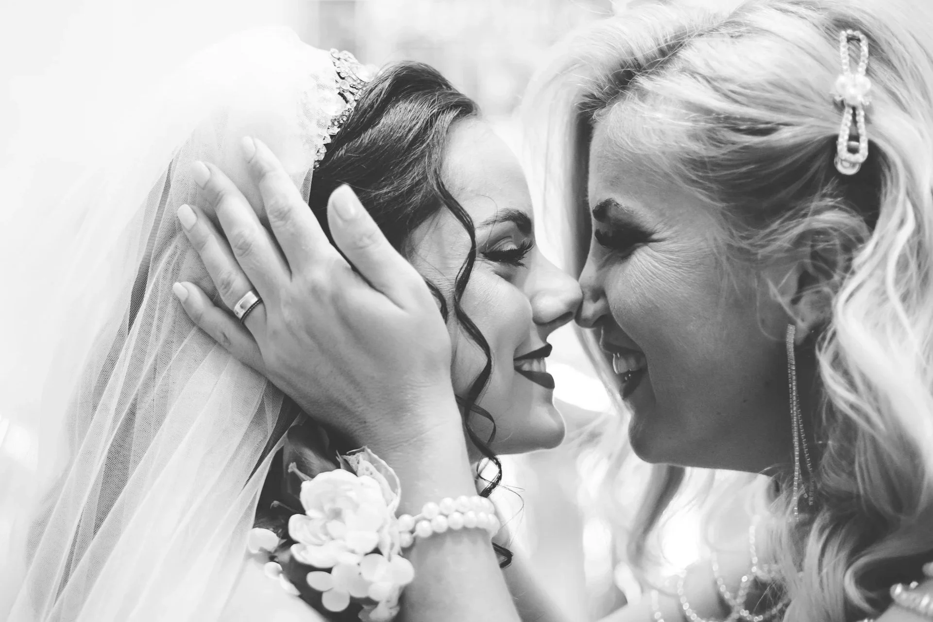 Two women with closed eyes gently touching their foreheads together, smiling in a close, intimate moment. One woman has darker, curled hair and a tiara, the other has lighter, blonde hair with decorative hair accessories. Both are dressed elegantly, and the woman with darker hair wears a strapless gown with floral accents.