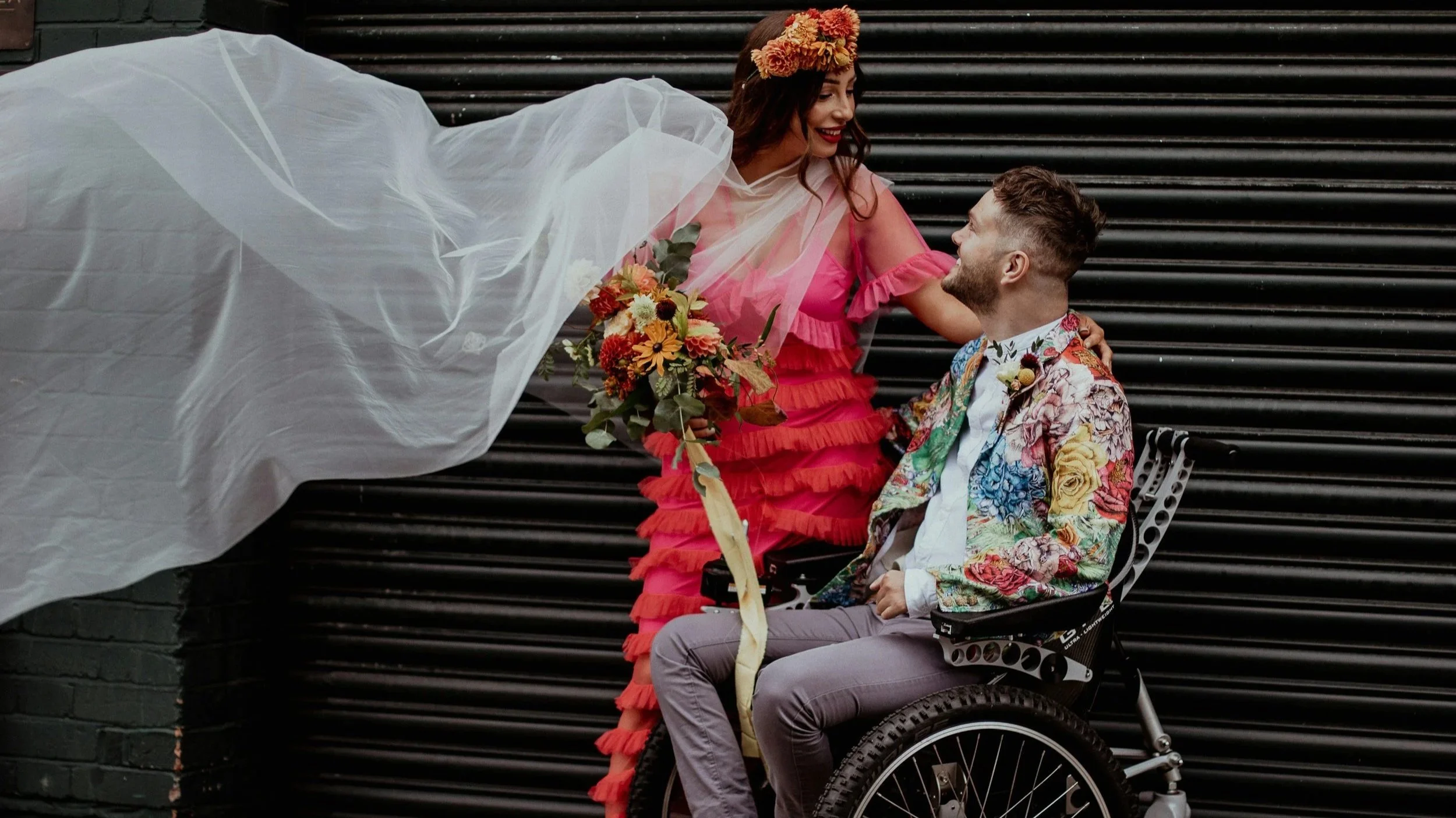 A woman in a pink dress with ruffles, wearing a flower crown, holding a bouquet of flowers, looking at a man in a floral suit jacket seated in a wheelchair, against a black corrugated metal background.
