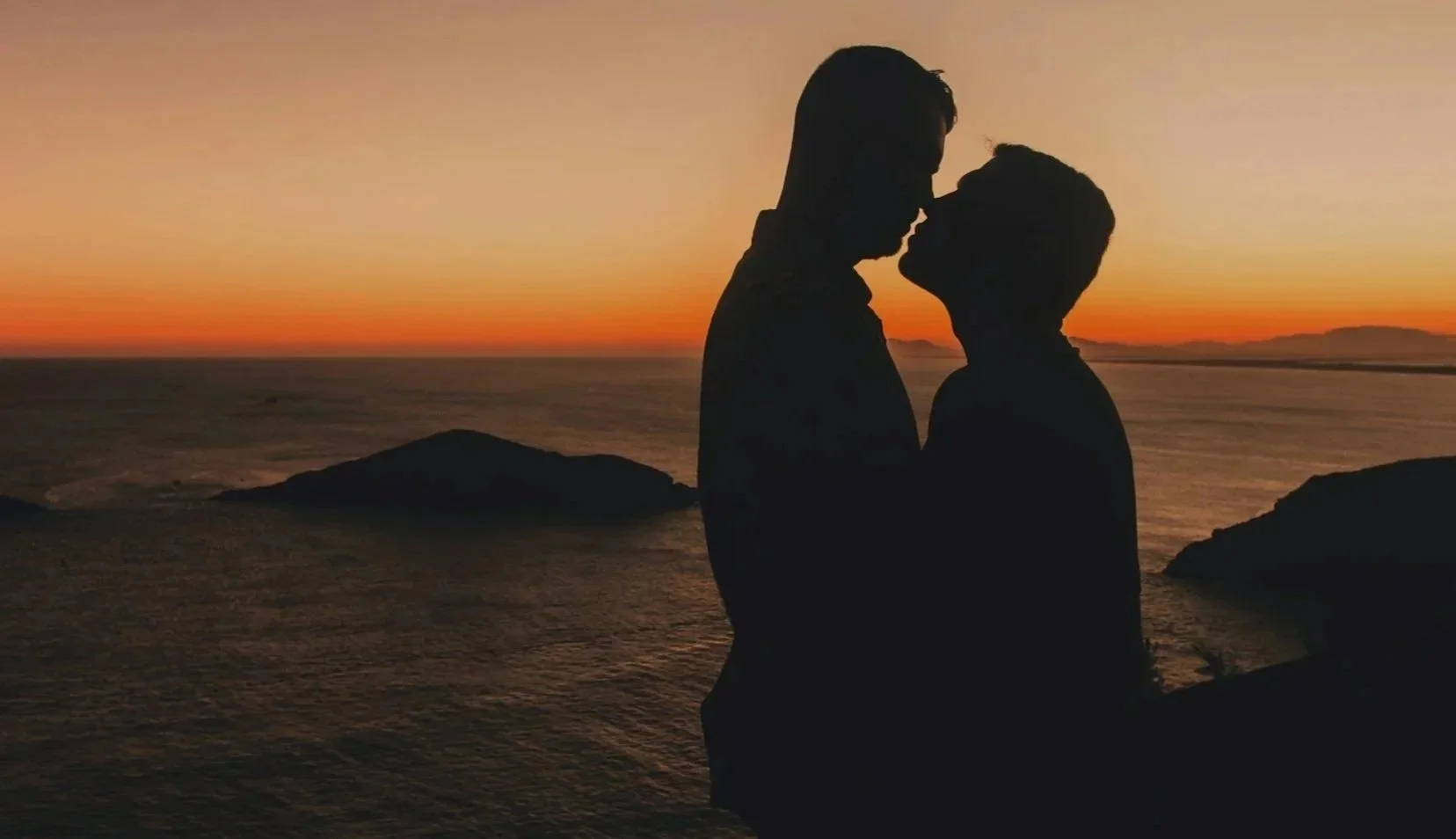Silhouettes of a couple about to kiss at sunset by the ocean, with rocks in the foreground and a colorful sky in the background.