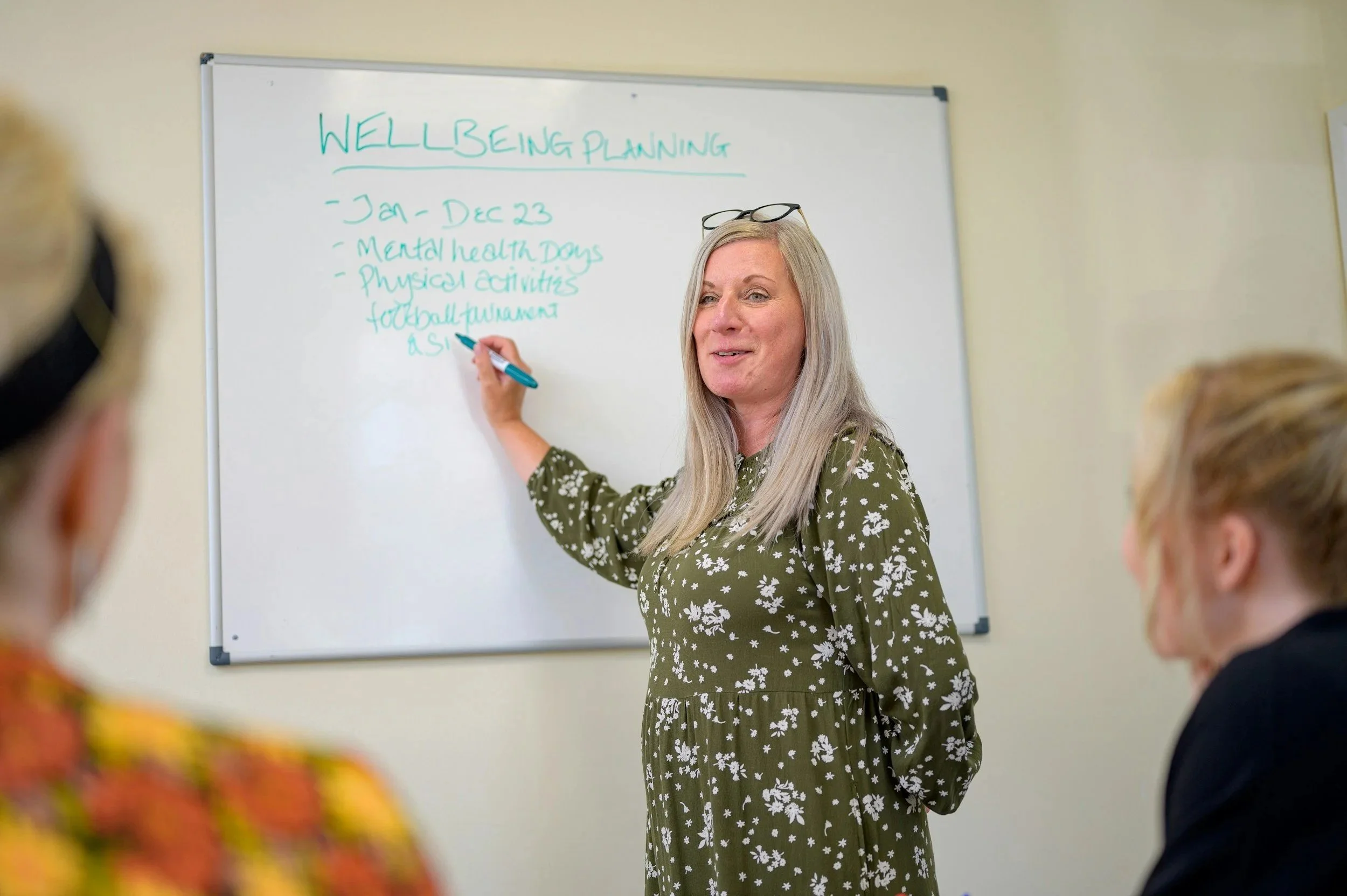 A woman with long gray hair and glasses resting on her head writes on a whiteboard in a classroom. The whiteboard has the words "WELL-BEING PLANNING" and a list, with students partially visible in the foreground.
