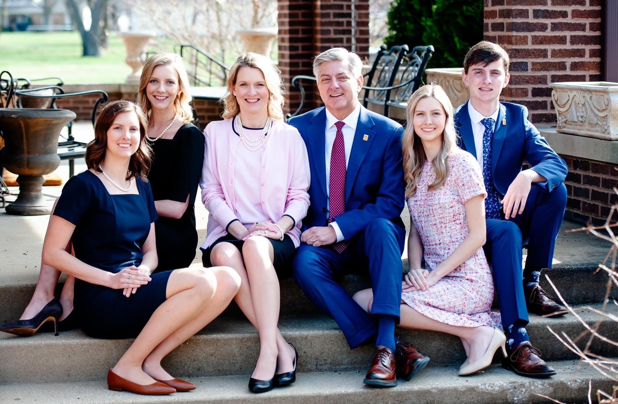 Group of six people sitting on steps outside, smiling, dressed in formal and semi-formal attire, with brick building and outdoor furniture in the background.