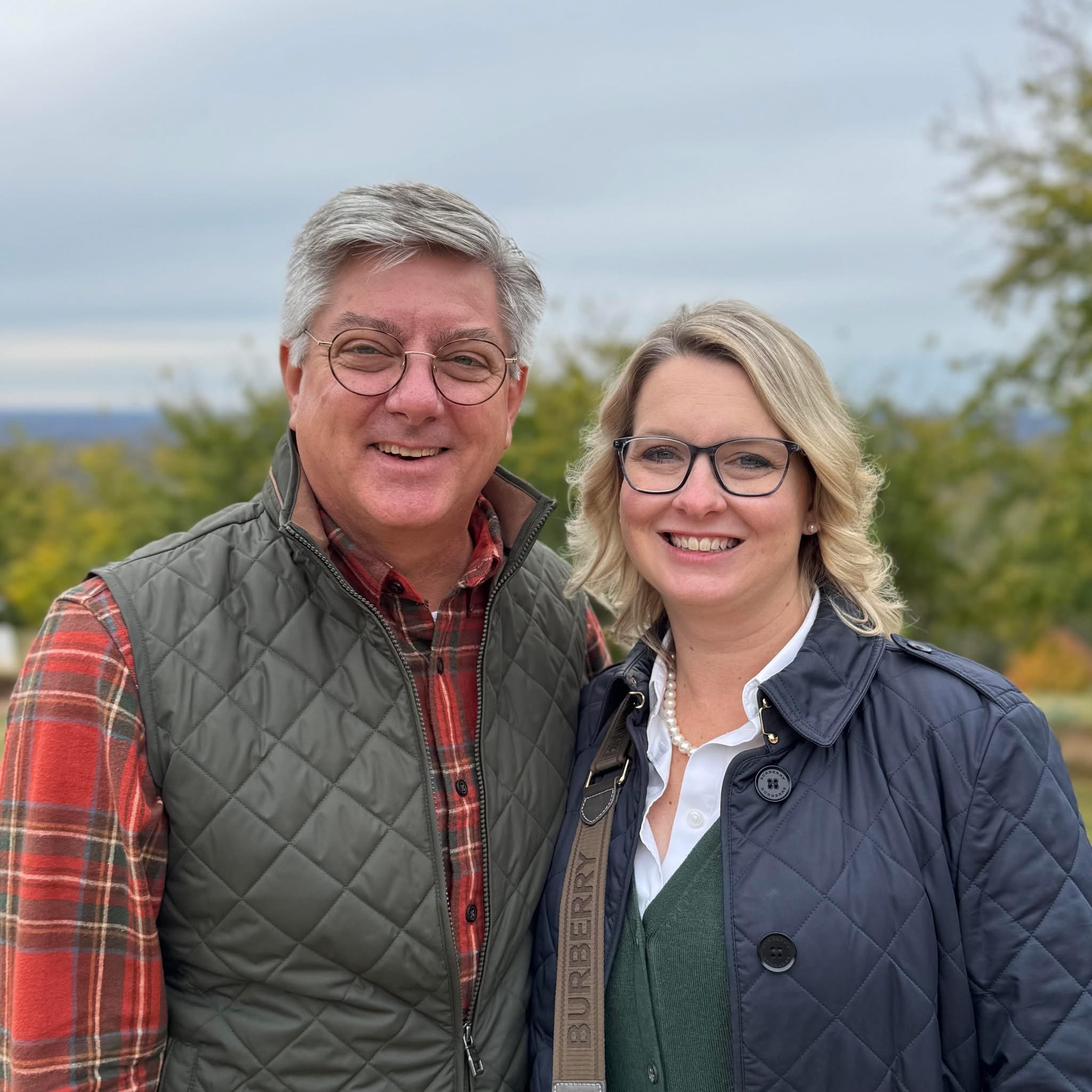 A middle-aged man and woman standing outdoors, both smiling at the camera with trees and a cloudy sky in the background.