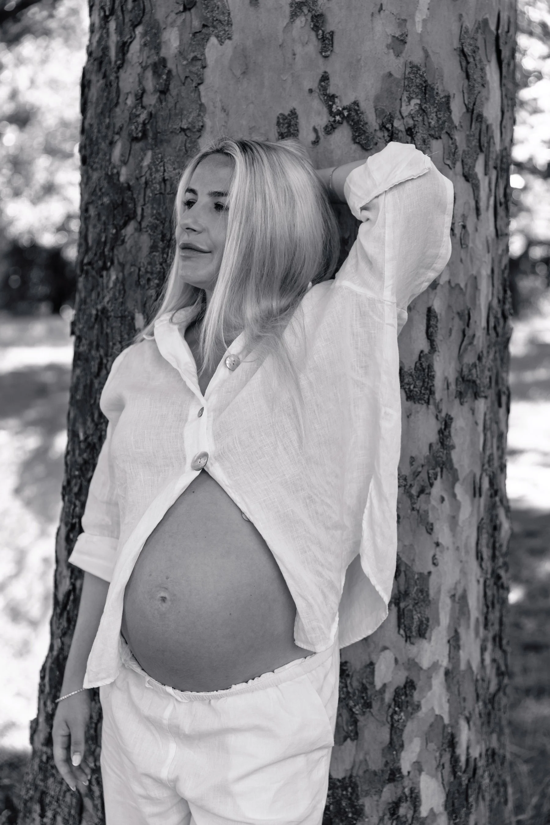 A pregnant first-time mum showing her bump dressed in a white linen shirt and trousers leaning on a tree during a maternity photoshoot in Wandsworth.