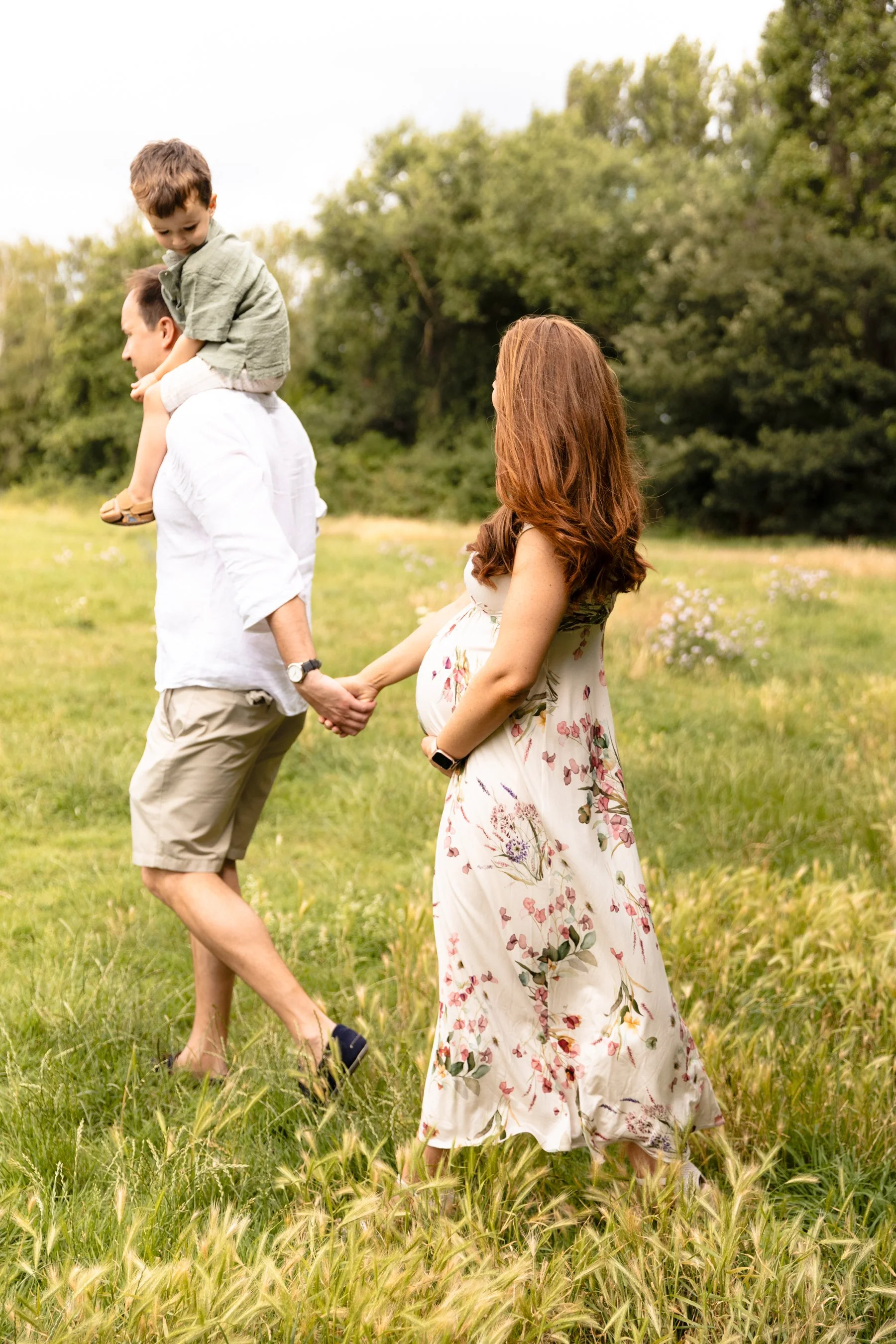 An expectant couple walking together outdoors in Wandsworth, with the father carrying their toddler son on his shoulders as the pregnant mother smiles.